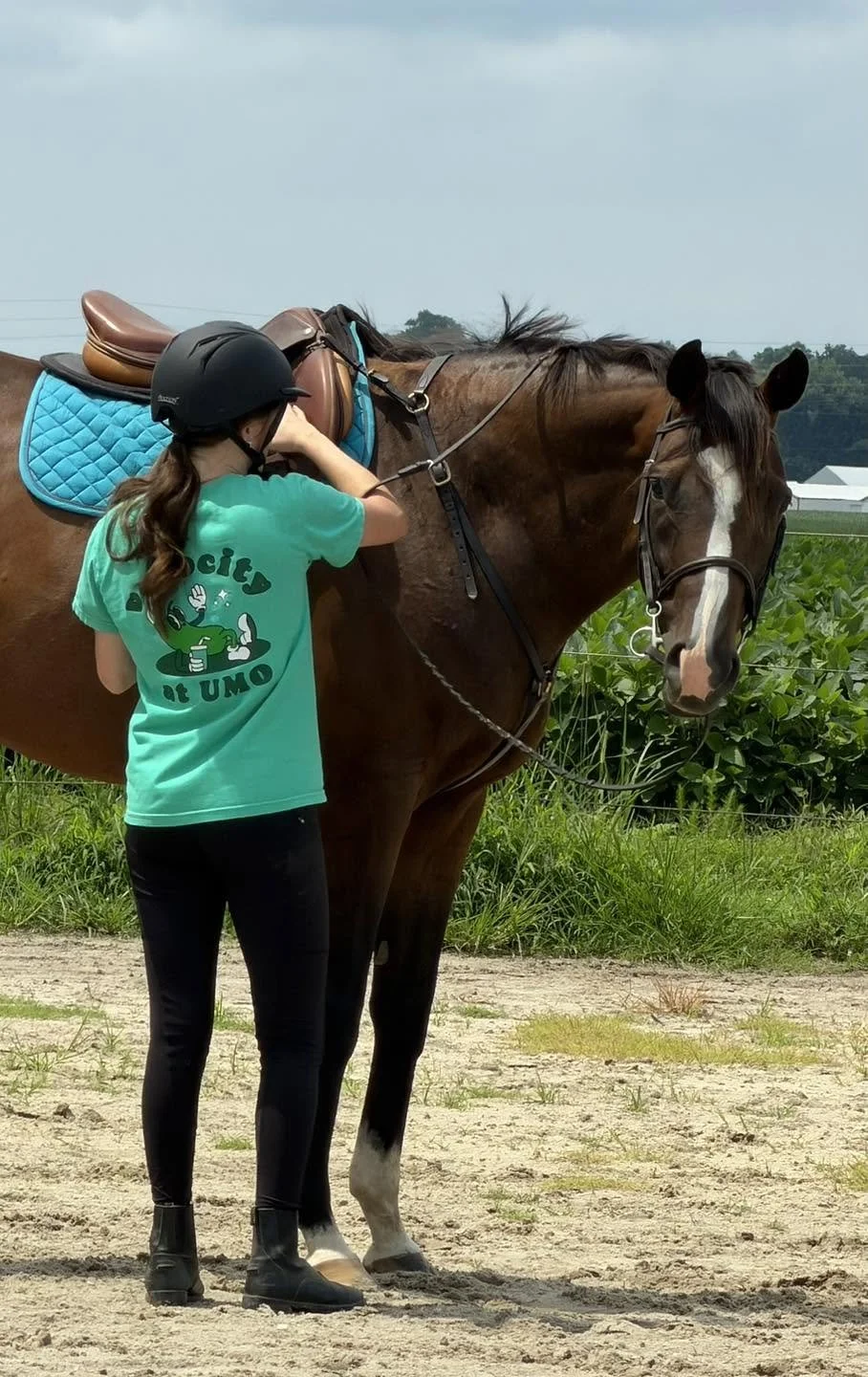 A girl wearing a helmet and a turquoise shirt is standing next to a brown horse with a blue saddle pad and a saddle. She appears to be adjusting the horse's bridle or equipment while preparing for riding outdoors on a dirt path with greenery and blue sky in the background.