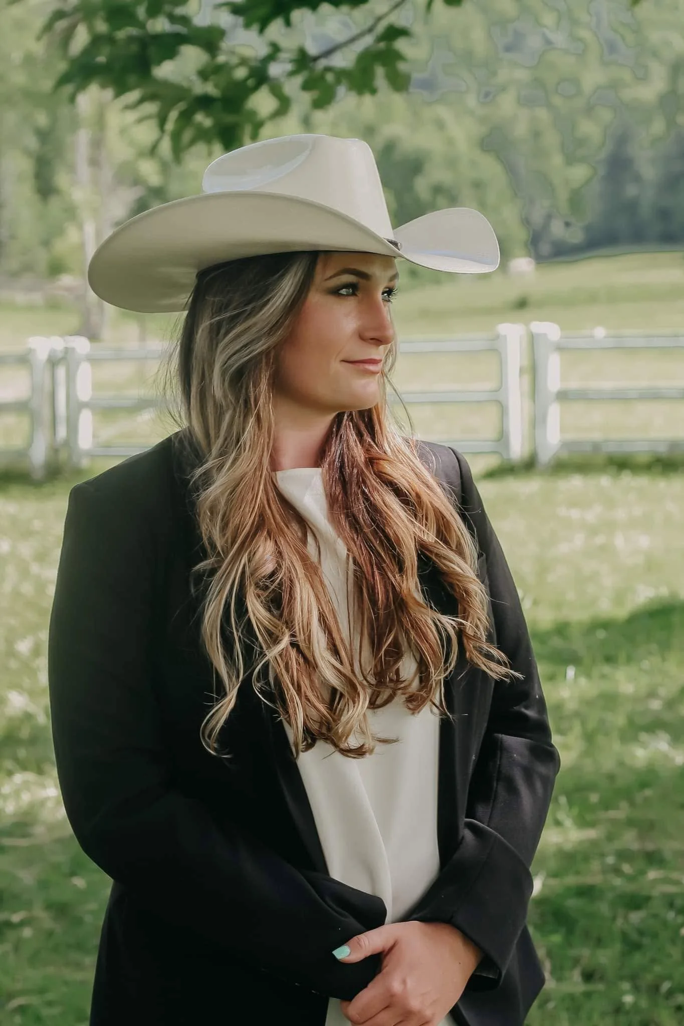 A woman with long wavy hair and light makeup, wearing a white cowboy hat and black blazer, stands outdoors in front of a white fence and green trees, smiling slightly and looking to her right.