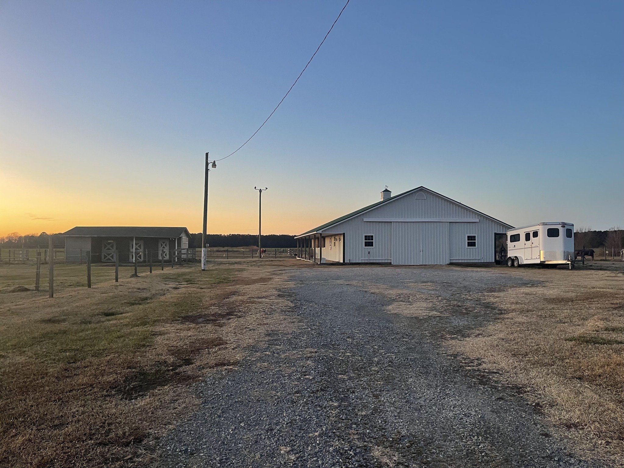 A rural scene at sunset featuring a white barn, a horse trailer, and horse pastures with fences under a blue sky.