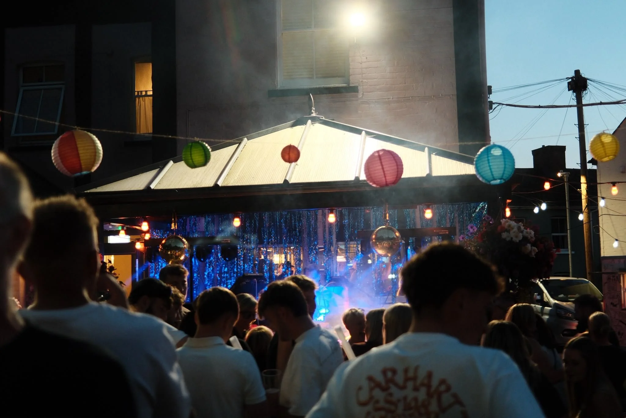 Crowd gathered outside during a festival or celebration, with colorful paper lanterns hanging overhead and a stage with blue lights in the background, under a clear sky.