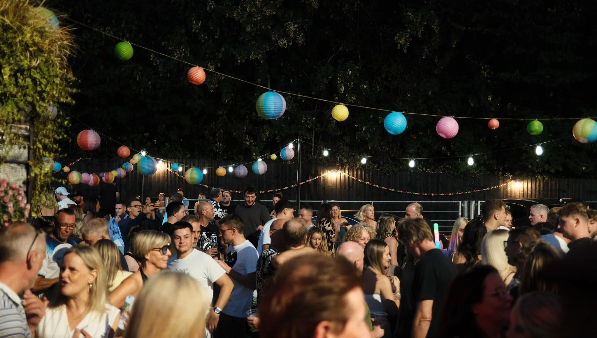 Crowd of people gathered outdoors at night, decorated with colorful paper lanterns and string lights.