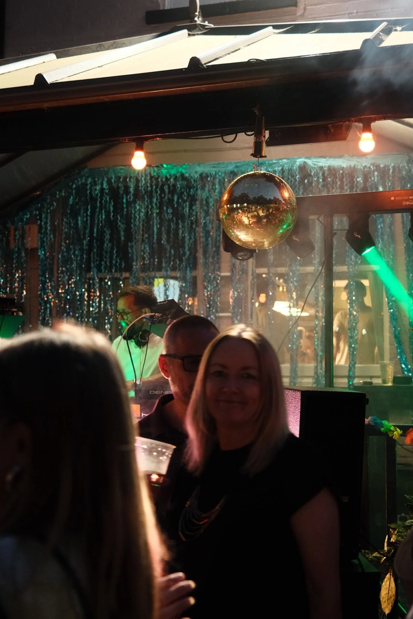 People dancing and socializing in a dimly lit party or nightclub with a disco ball, string lights, and colorful lighting.