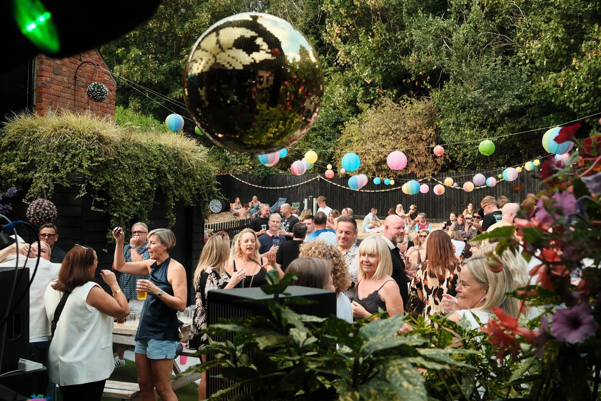 A crowded outdoor party with people socializing under colorful paper lanterns hung on strings in a lush backyard setting. The image is viewed from behind a reflective disco ball.