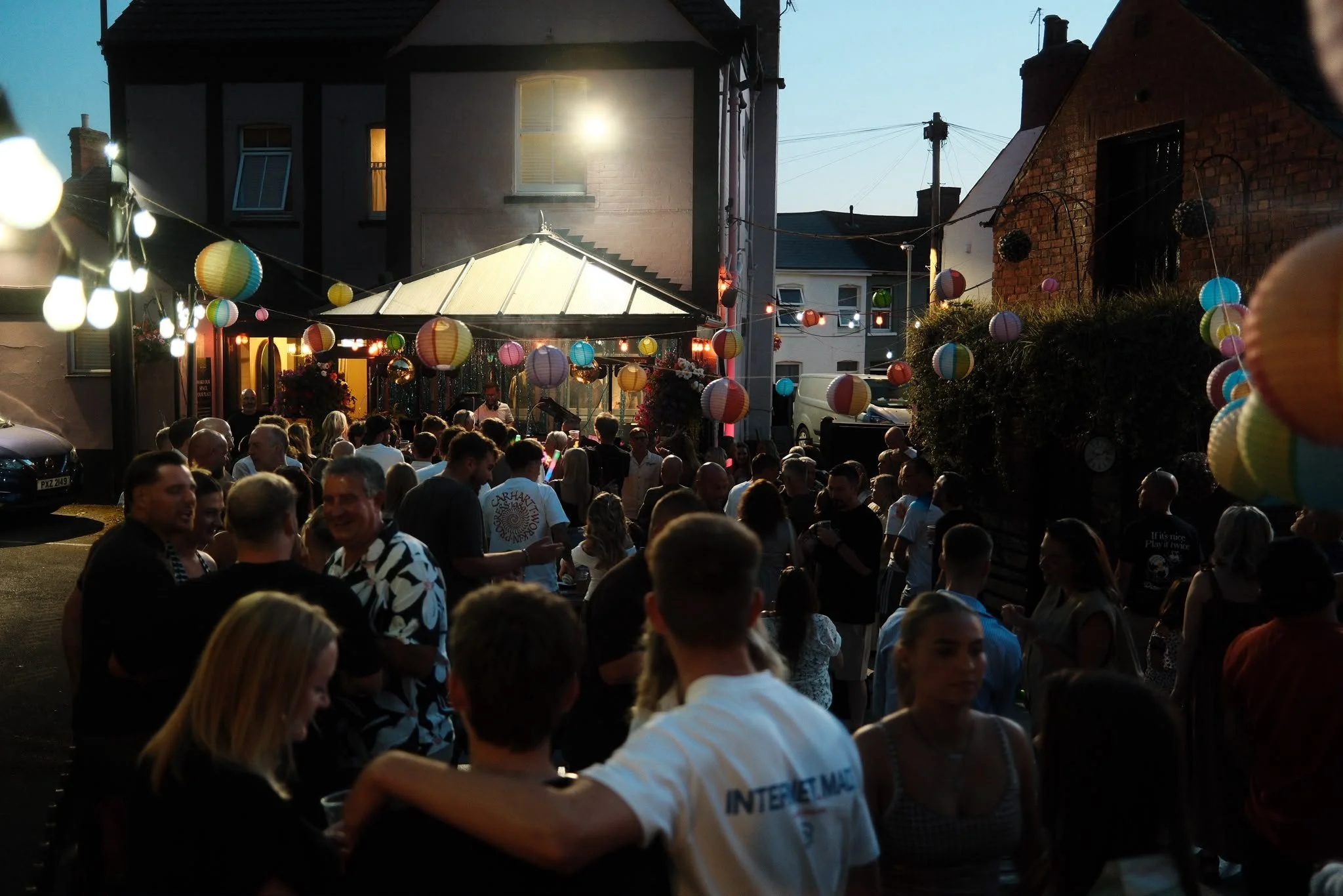 A lively outdoor gathering during the evening with many people socializing, decorated with colorful paper lanterns and string lights, set against buildings with a mix of older brick and modern architecture.