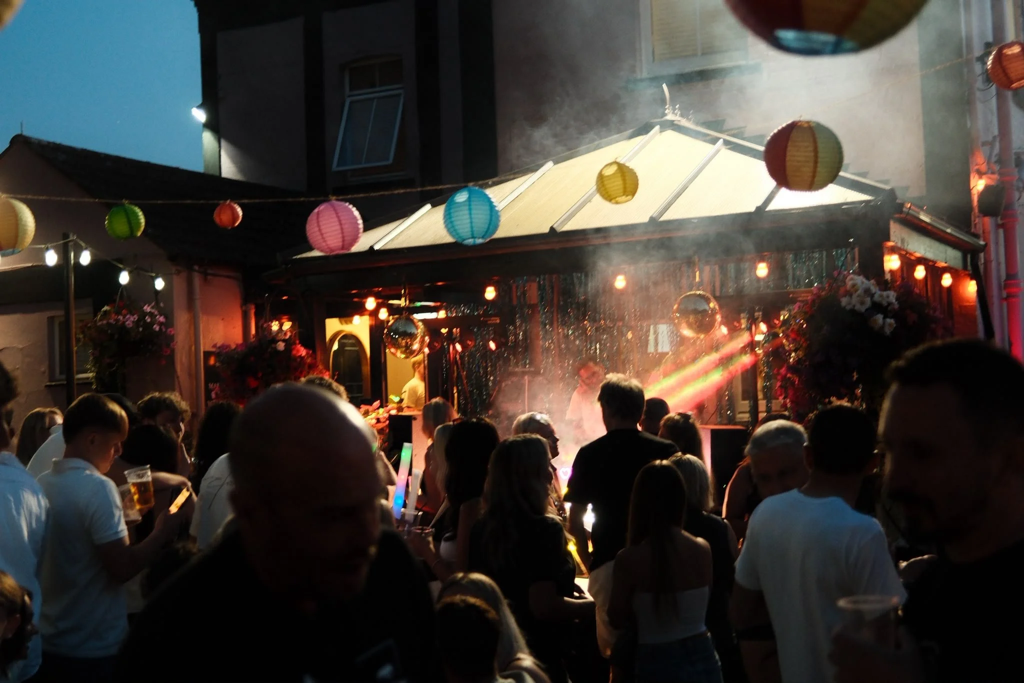 A crowded outdoor party at night with colorful paper lanterns hanging overhead, people holding drinks, and a band performing on a stage with bright lights.