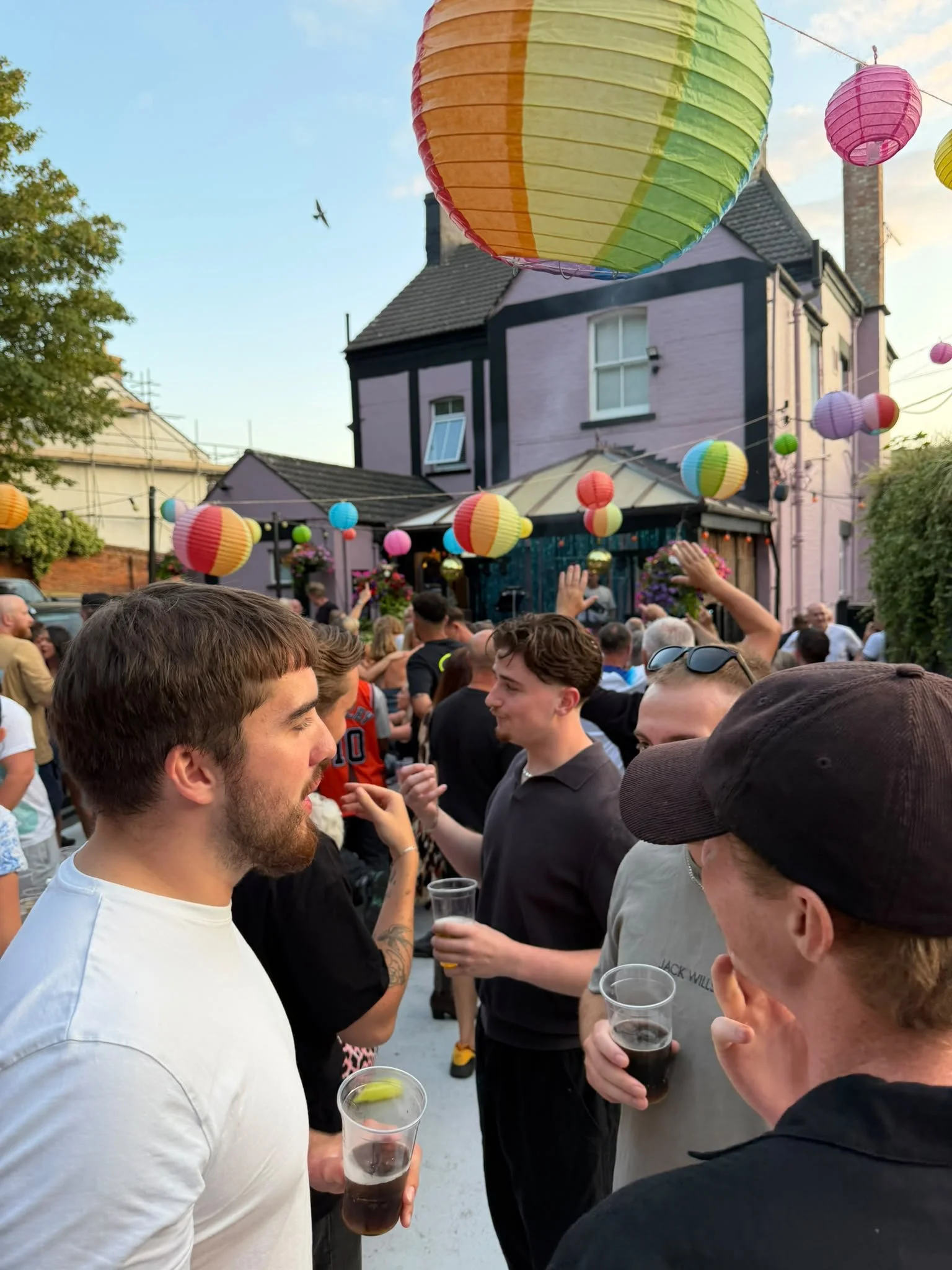 A group of people gathered outdoors during a festival or celebration, with colorful paper lanterns hanging overhead. People are holding drinks and engaging in conversations near a building with purple and black facades.