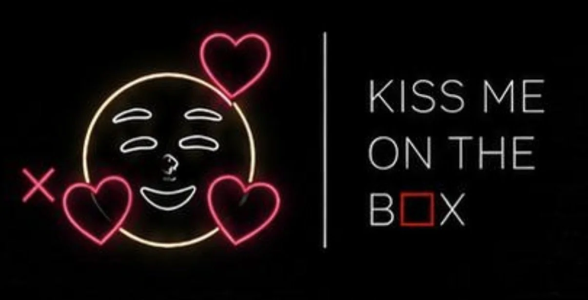 Neon sign with a smiling face surrounded by three pink hearts, and text 'Kiss me on the box' with the words 'box' having a red square in place of the letter 'o'.