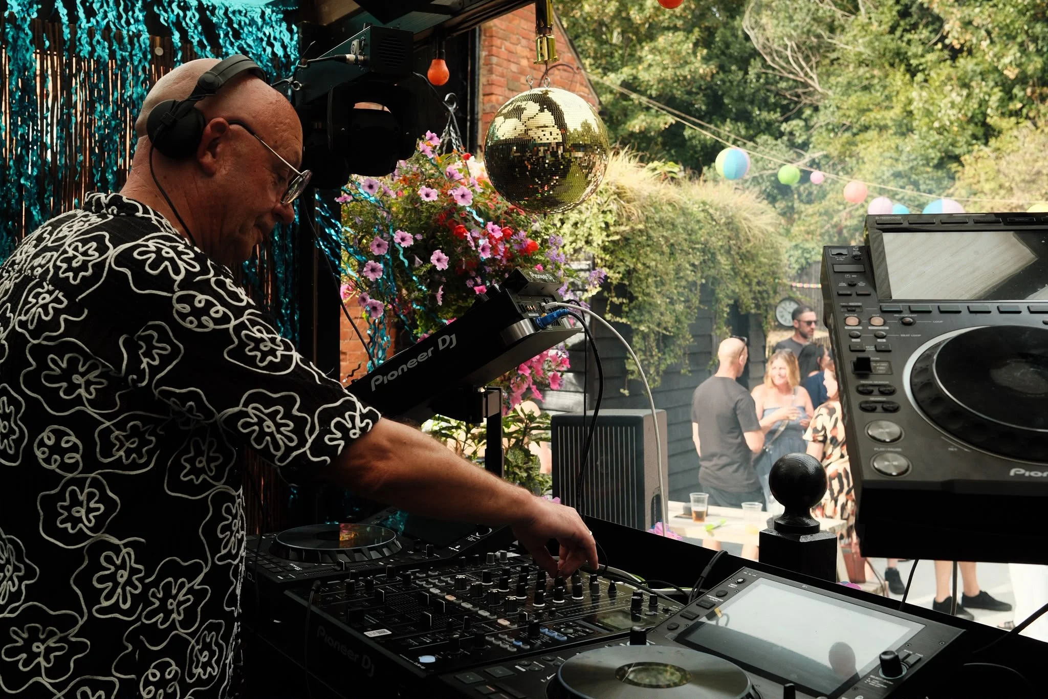 A DJ wearing glasses and a patterned black and white shirt plays music at an outdoor event. He is using a Pioneer DJ controller, with a large mixing deck and a screen. A gold disco ball and colored paper lanterns hang above. There are pink flowers and green trees in the background, along with people mingling and enjoying the event.