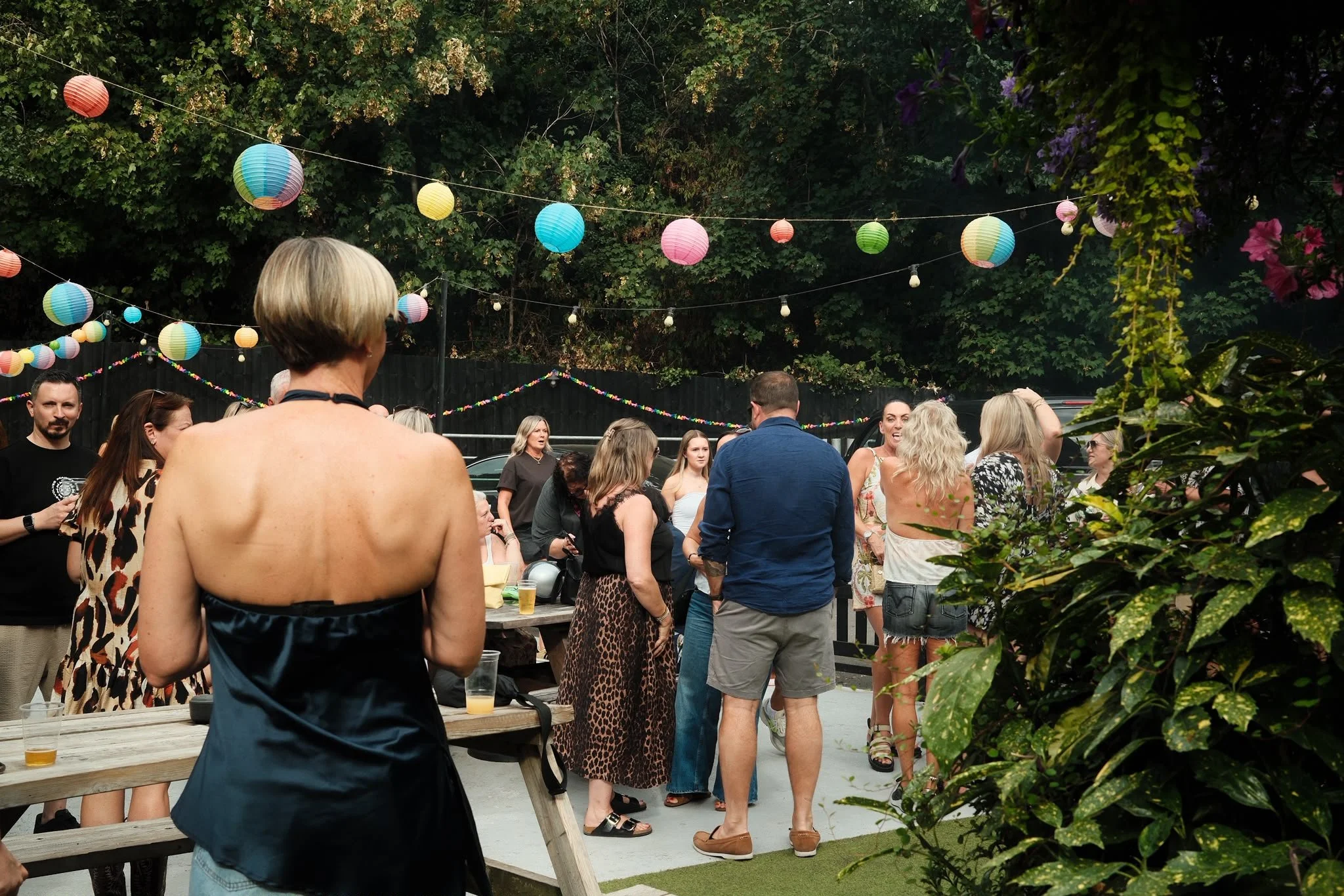 People gathering outdoors for a social event with colorful paper lanterns hanging above, surrounded by trees and greenery.