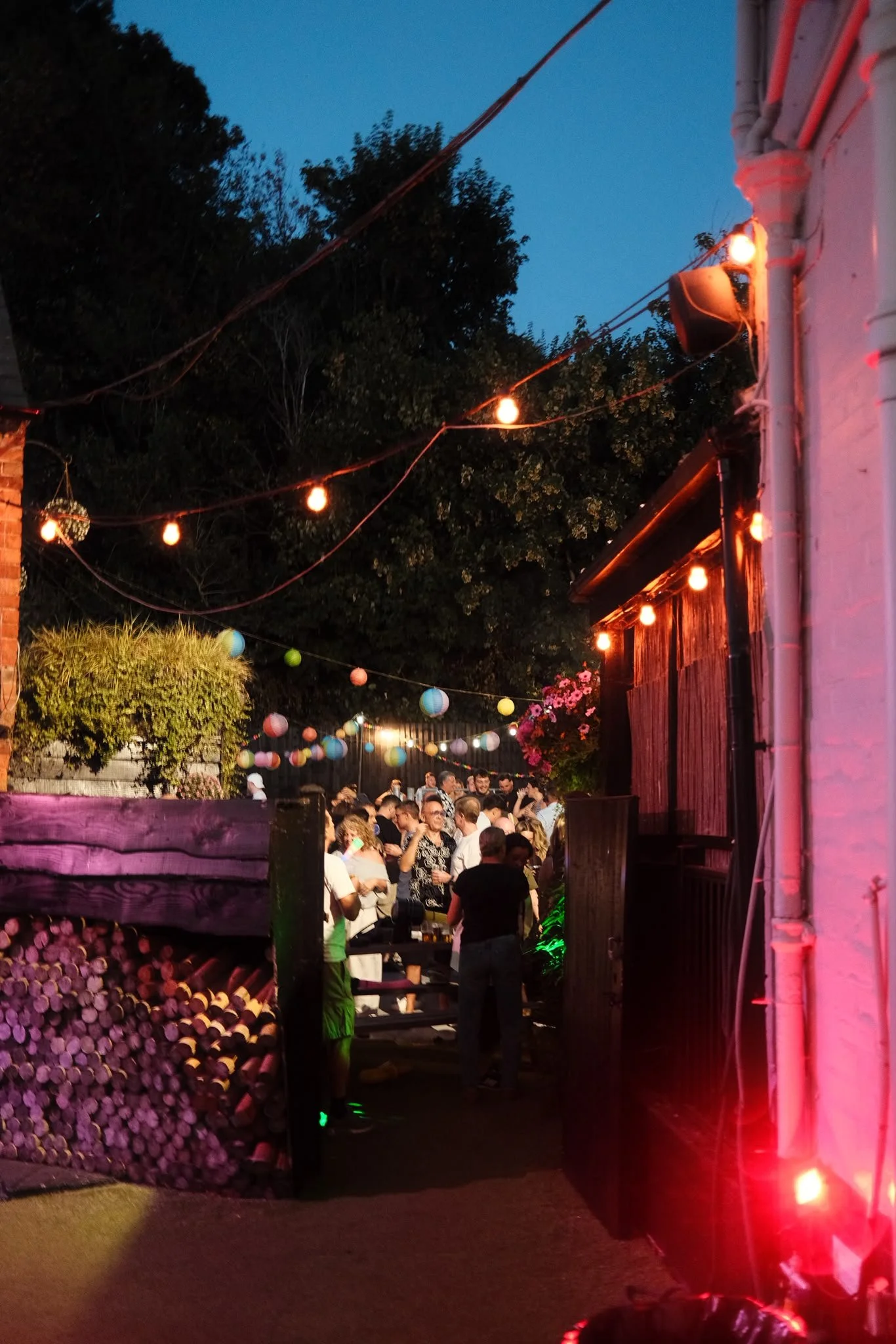 Nighttime outdoor gathering with string lights, people socializing, and a wooden wall with stacked firewood on the left. The scene is illuminated with warm lights, creating a festive atmosphere.