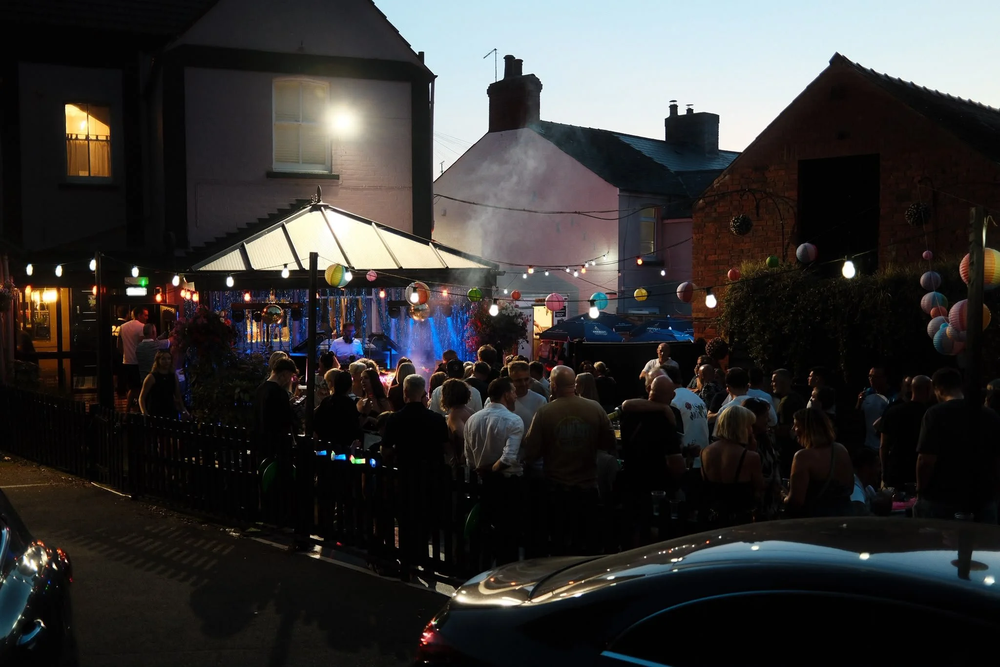 A lively outdoor gathering at dusk with people socializing under string lights, a small stage with a live band, and houses with lit windows in the background.