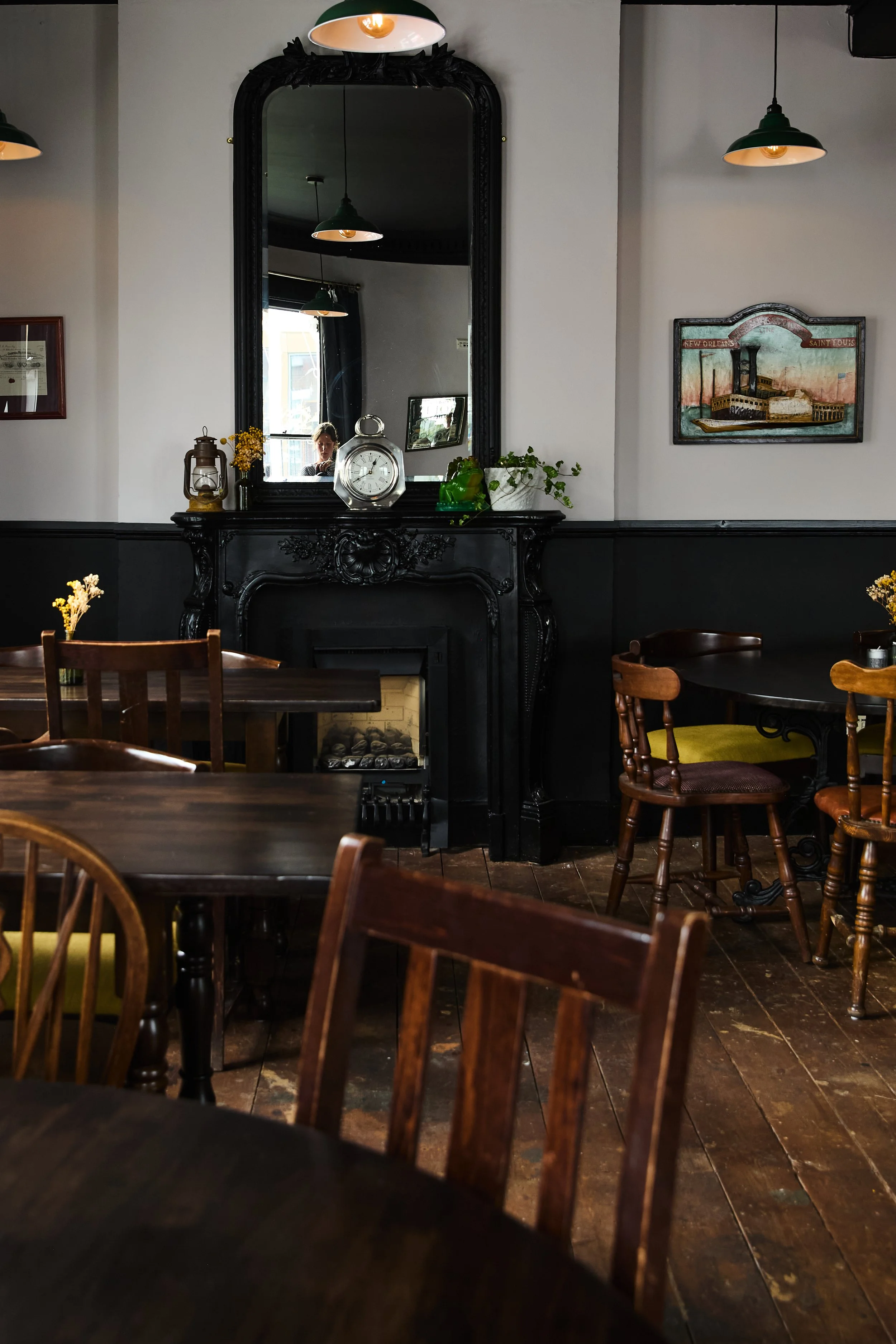 Interior of a cozy restaurant with wooden tables and chairs, a decorative black fireplace mantle with a mirror above, and framed artwork on the wall. The atmosphere is warm and inviting.