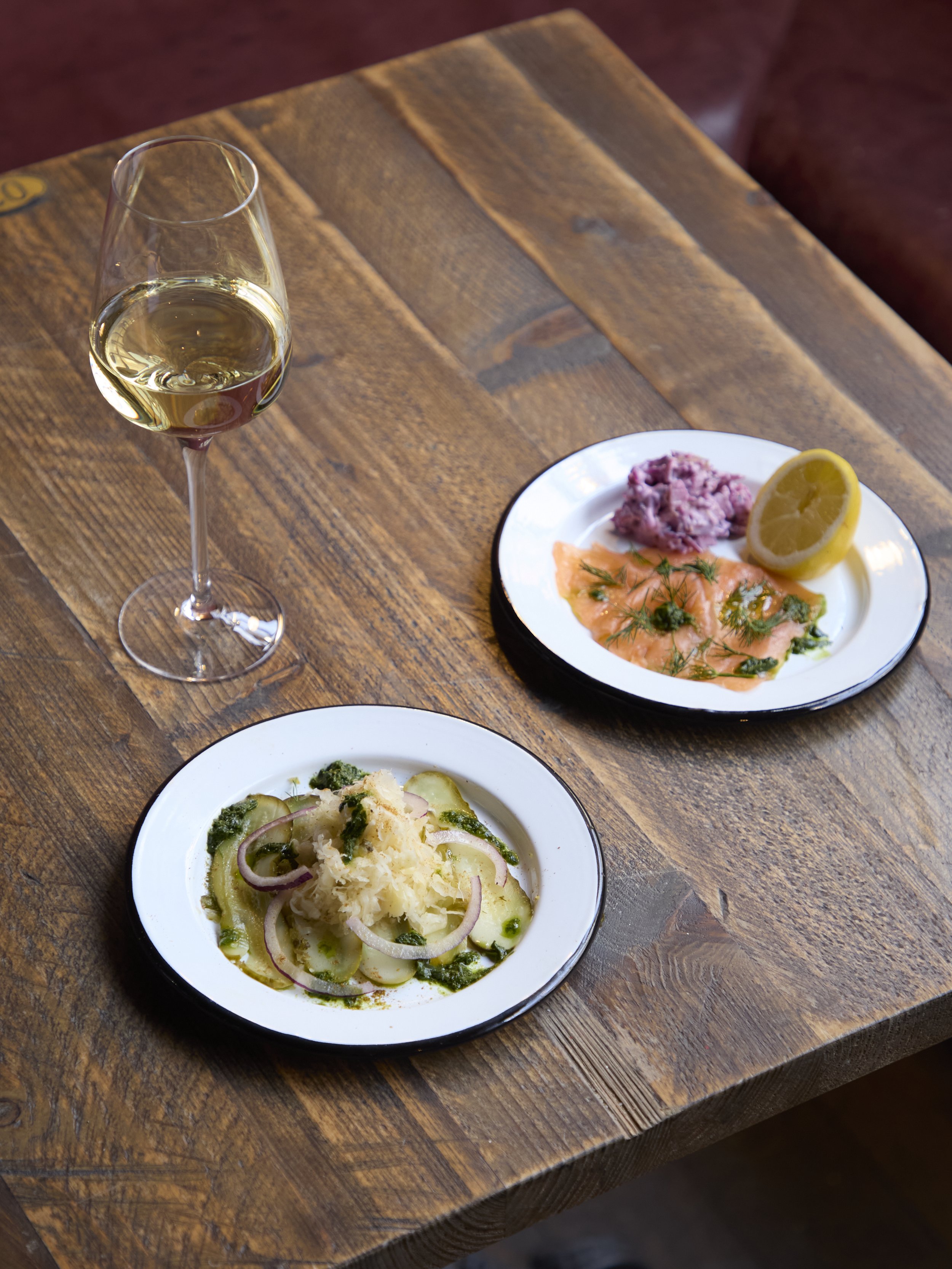 A wooden table with a glass of white wine, a plate of smoked salmon with herbs and lemon, and a plate of potato salad with onions and herbs.