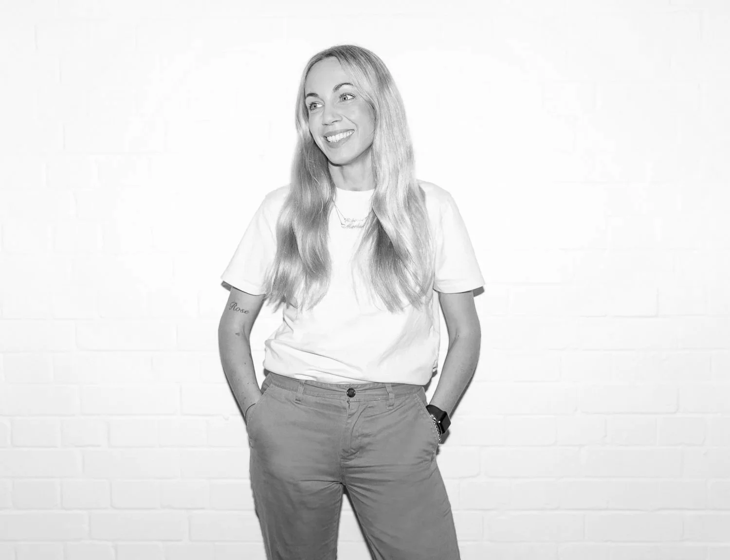 A woman with long blonde hair smiling, wearing a white t-shirt and pants, standing against a white brick wall.