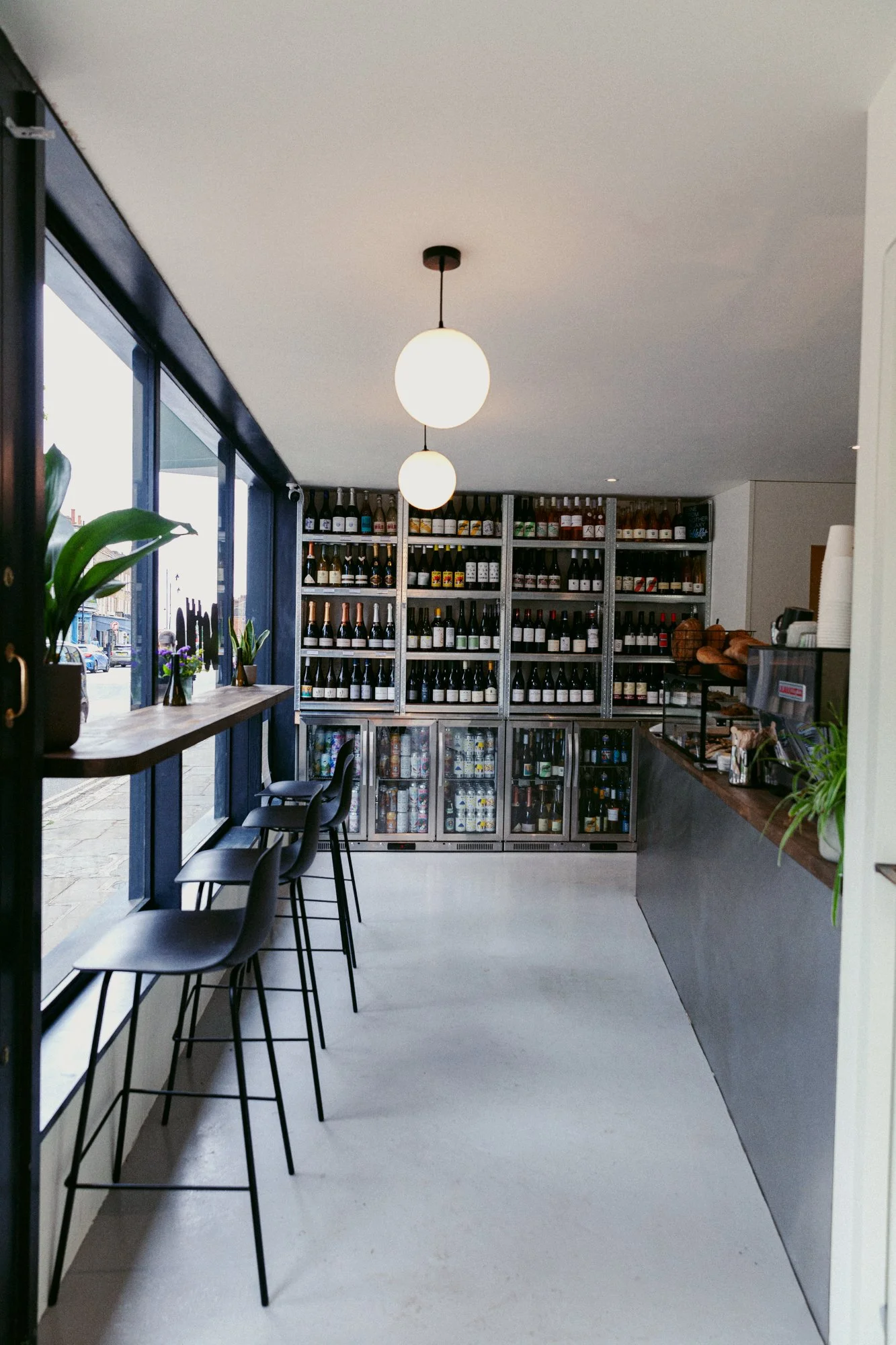 Interior of a modern restaurant or cafe with large windows, black chairs, a high bar-style table with plants in vases, and a wall of shelves filled with wine bottles. Pendant lighting hangs from the ceiling.