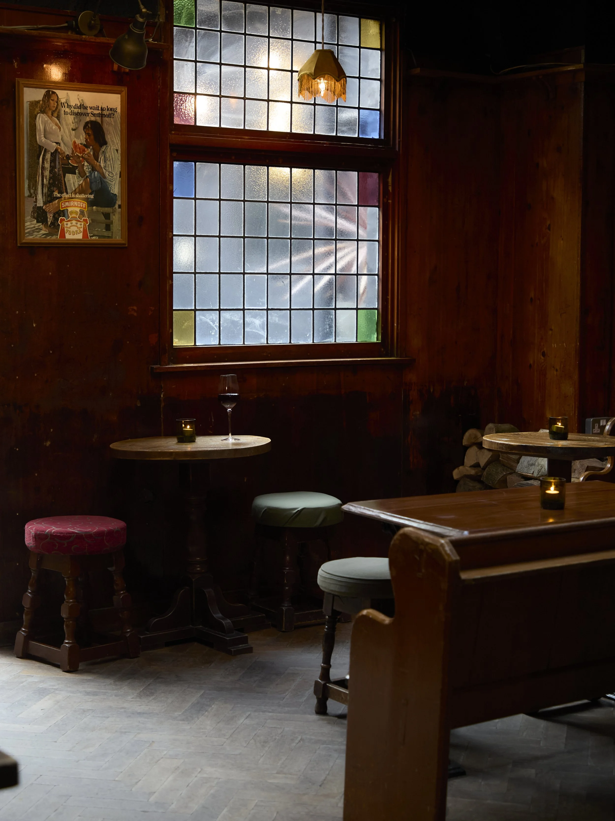 Inside a dimly lit rustic restaurant or café with wooden walls and furniture, a table with a single glass of red wine, small candles, and a stack of firewood in the corner. A large stained-glass window with rain droplets and sunlight creating a glow,