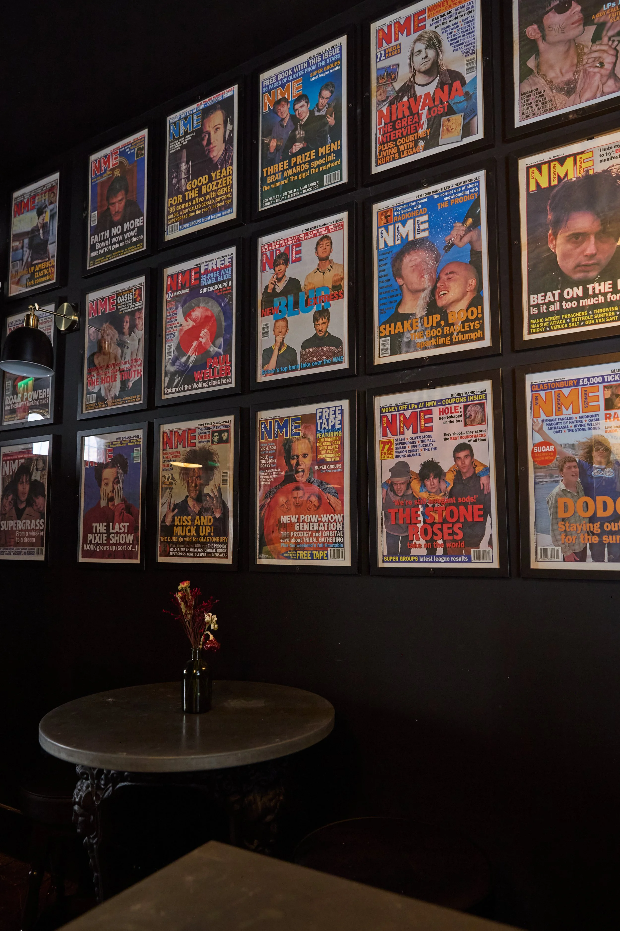 Wall decorated with framed magazine covers, mostly NME, featuring various musicians and bands, a round table with a small vase of dried flowers, and a black wall in the background.
