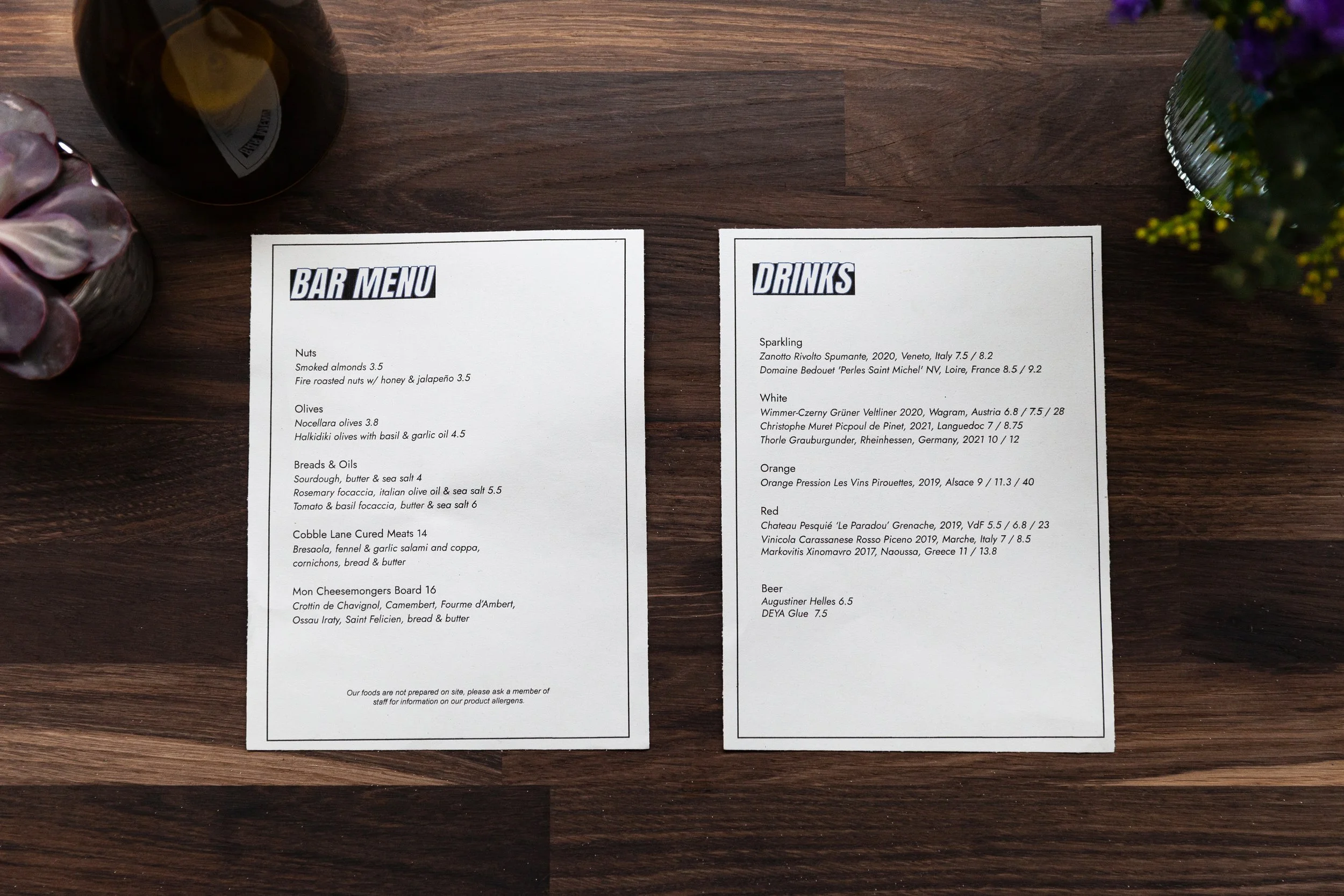 Two printed menus, one titled 'Bar Menu' listing various nuts, olives, breads, oils, cheese, and meats, and the other titled 'Drinks' listing different wines, beers, and non-alcoholic beverages, placed on a wooden table with a flower arrangement and 