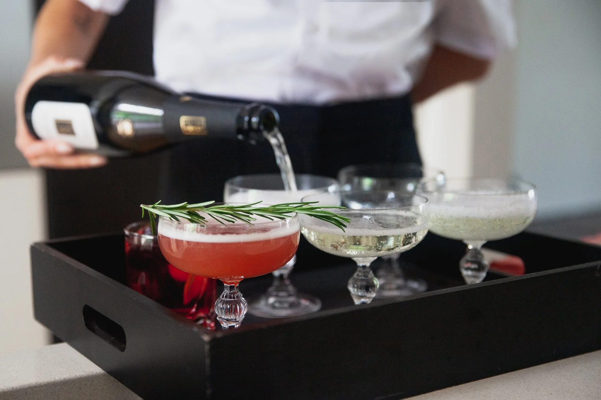 A person pours sparkling wine into decorative cocktail glasses on a black serving tray, with a sprig of rosemary garnish in one glass.