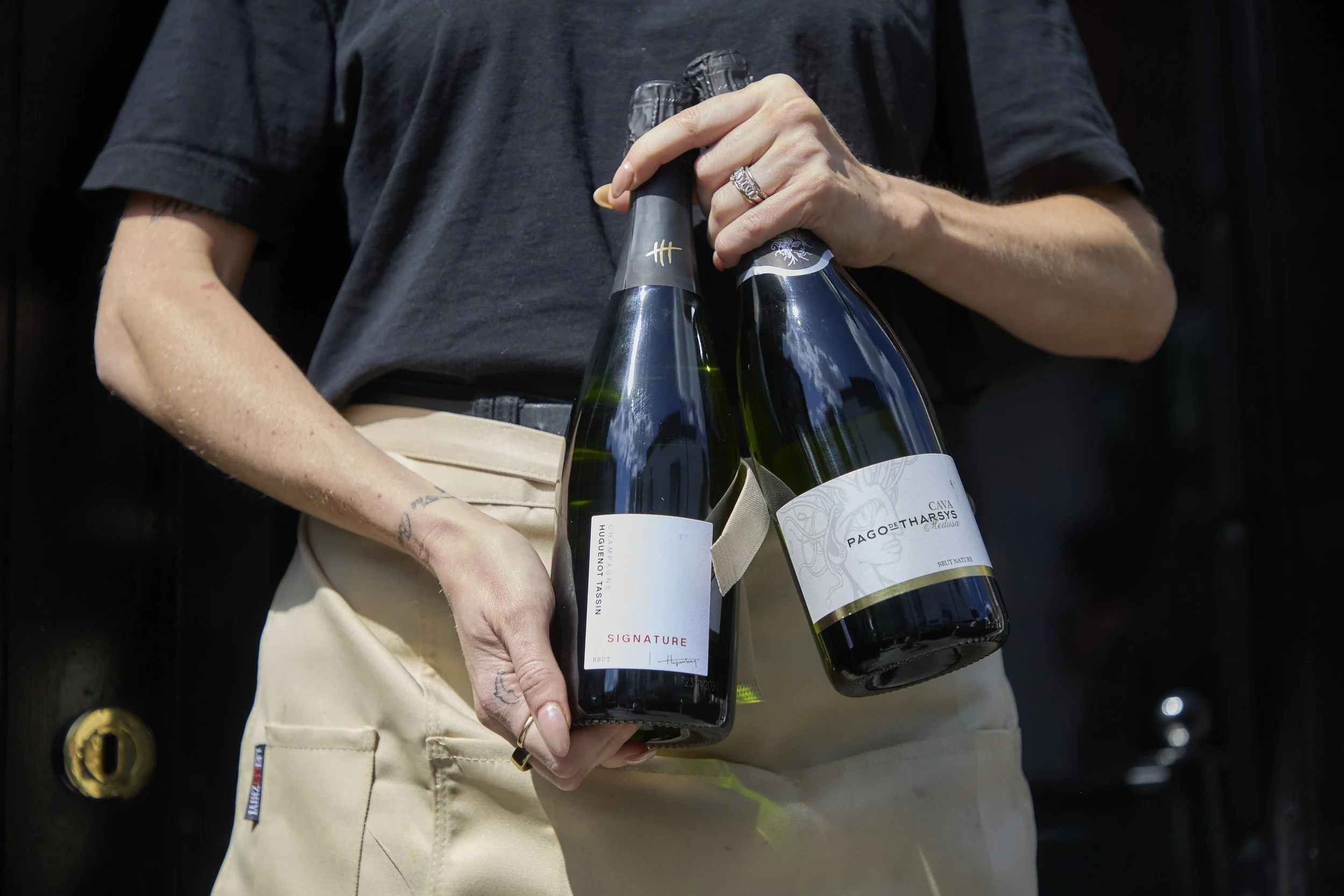 Person in black shirt holding three bottles of sparkling wine or champagne in their hand, standing outdoors.
