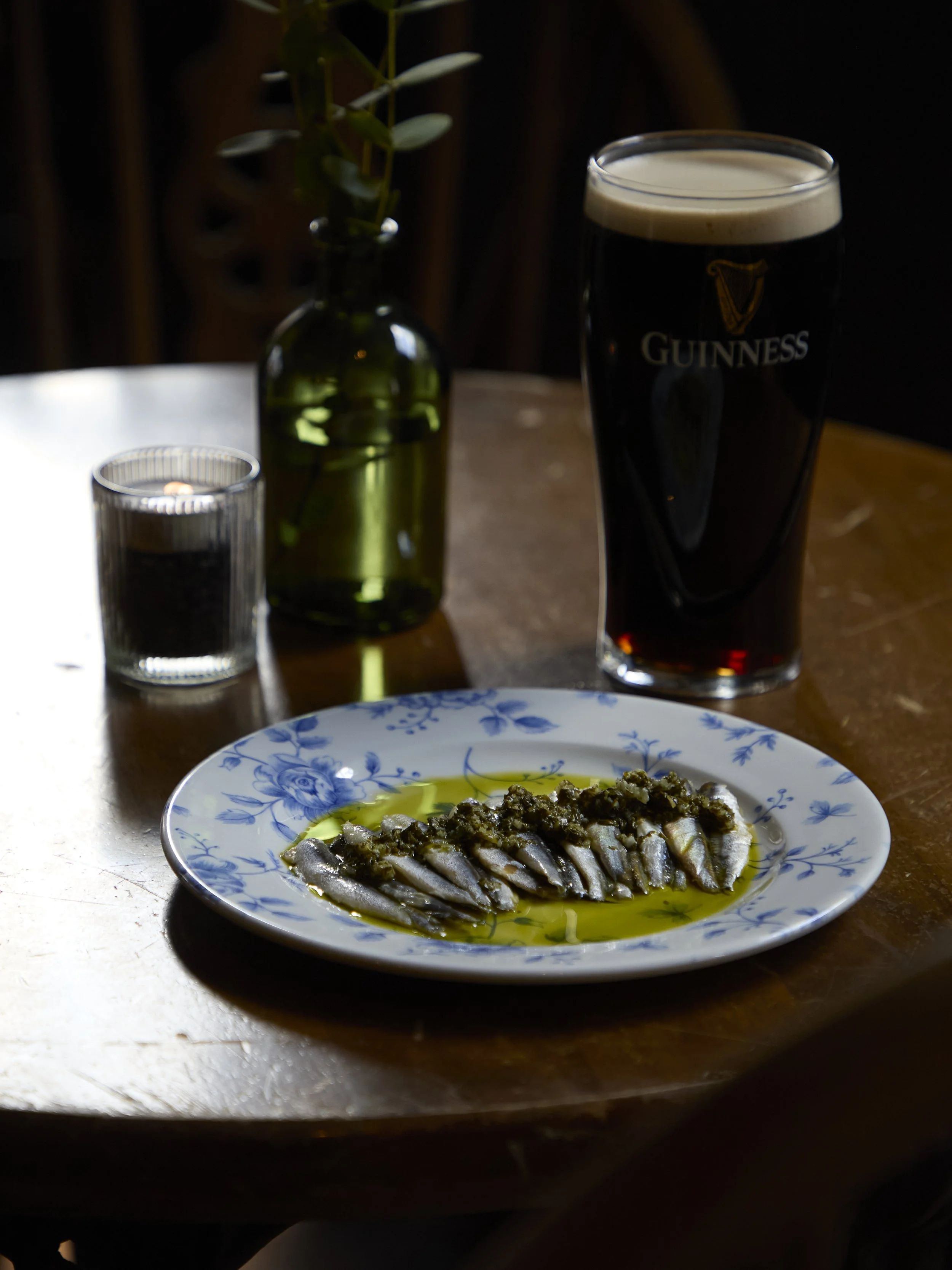 A plate of anchovies topped with herbs and drizzled with olive oil. In the background, there is a Guinness beer glass, a small candle, and a green glass bottle with a plant inside.