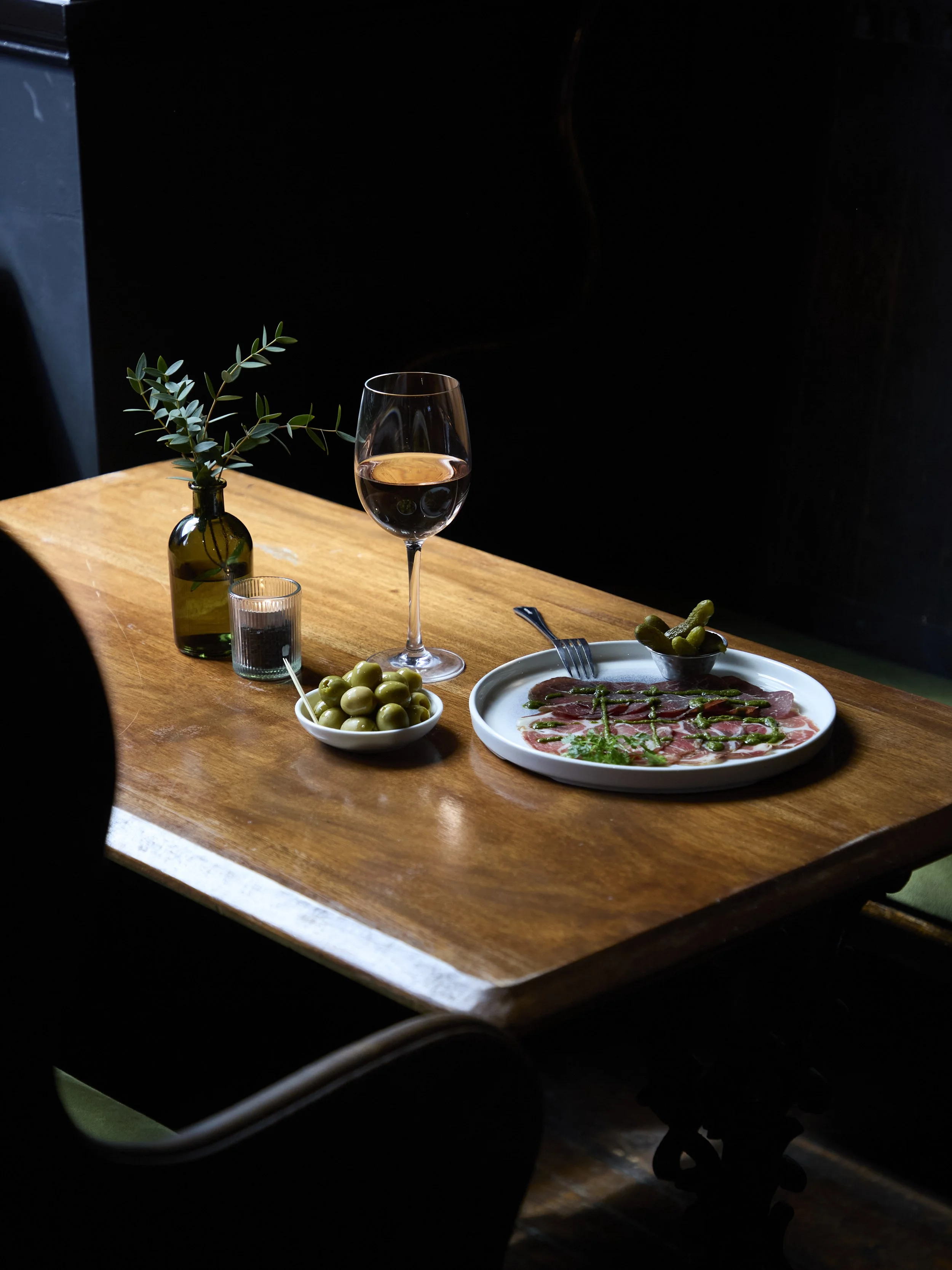 A table with a glass of rosé wine, a small vase with green leaves, a small candle holder, a bowl of green olives with a toothpick, a plate of cured meat with green sauce, and a fork, set against a dark background.