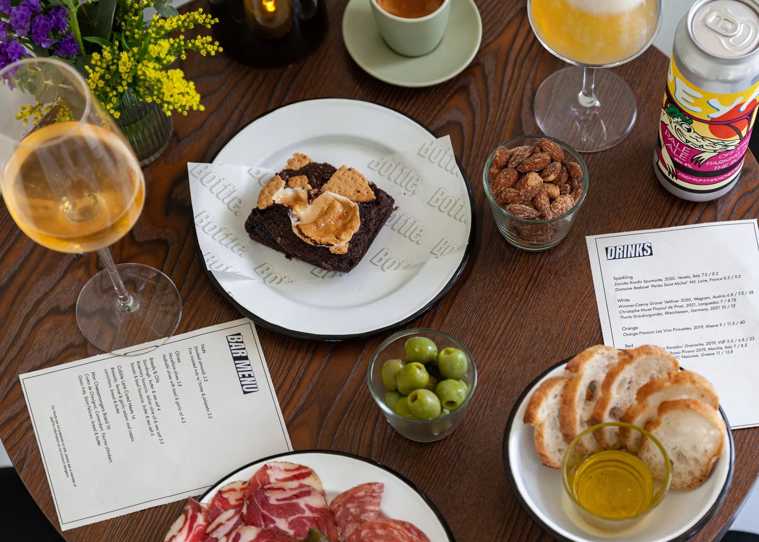 A table with various food and drink items, including a slice of brownie with ice cream, glasses of white wine, a bowl of pistachios, a bowl of almonds, green grapes, a beer can, a cup of tea or coffee, a flower arrangement, and menus.