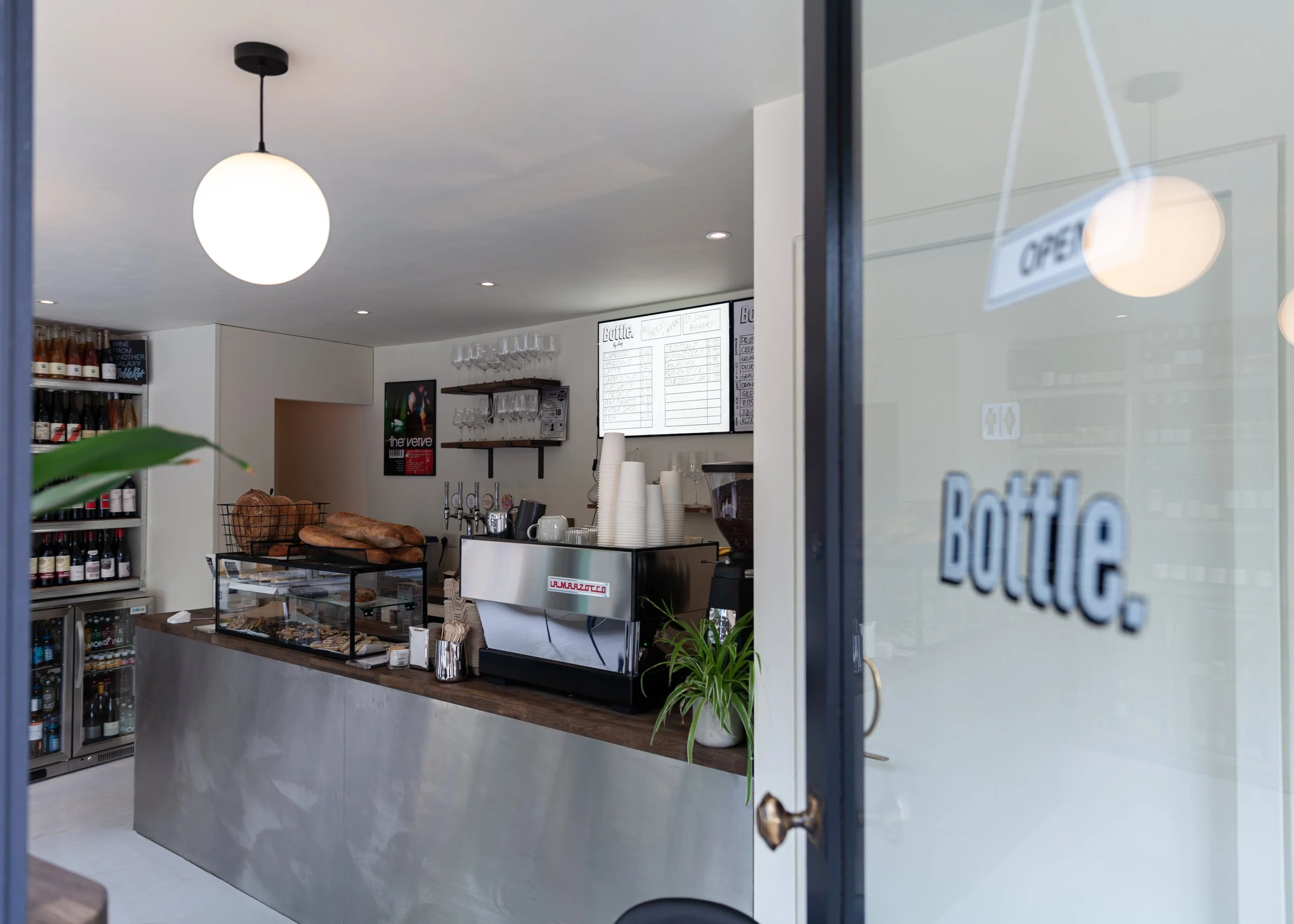Interior of a coffee shop or café with a counter displaying baked goods, a menu on the wall, wine bottles on shelves, and a door with an 'OPEN' sign.