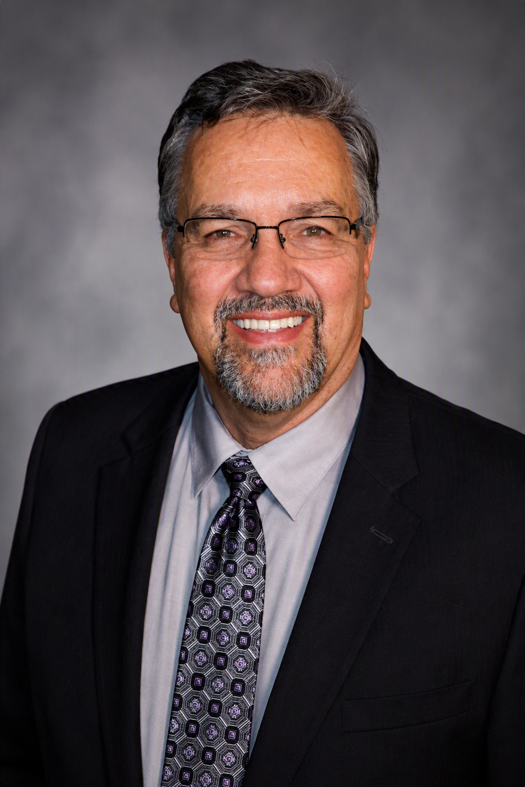 Portrait of a smiling middle-aged man wearing glasses, a suit, light shirt, and a patterned tie, with a dark gray background.