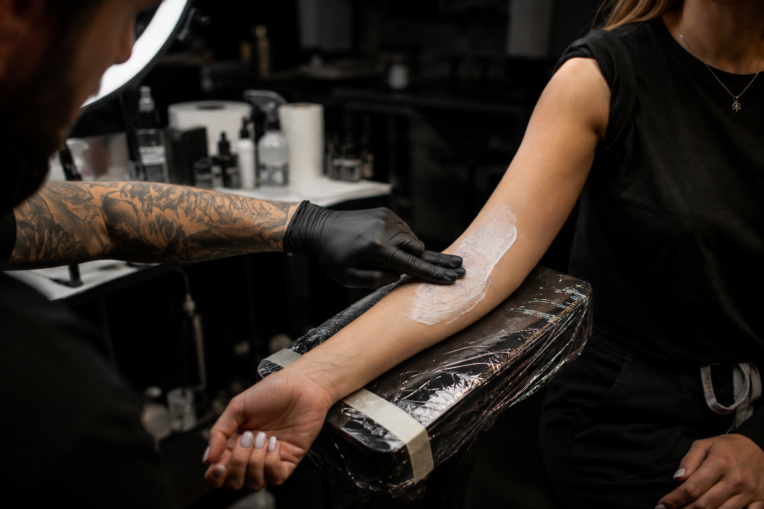 Tattoo artist applying a tattoo stencil to a female client's forearm in a tattoo studio.