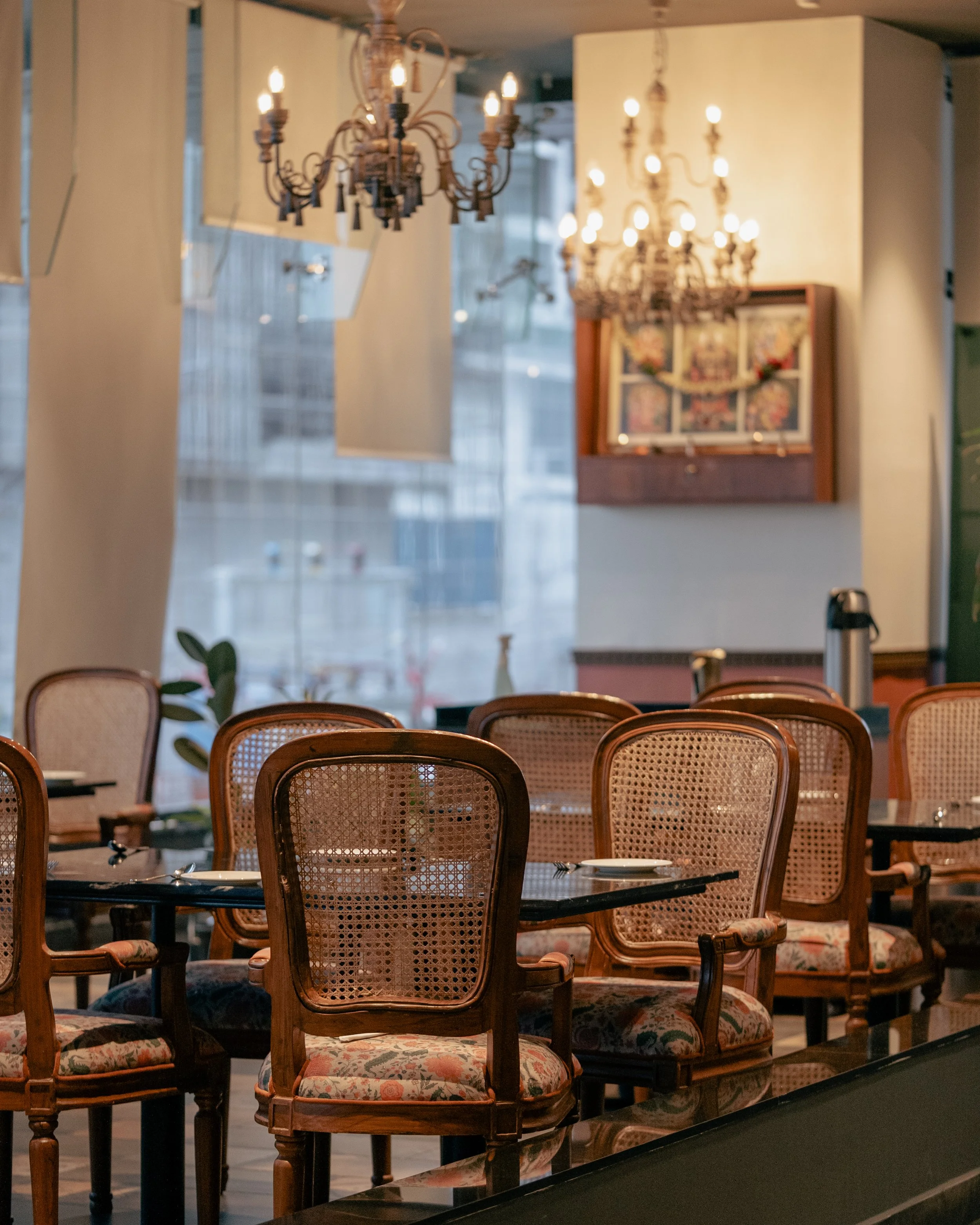 A dining area with multiple wooden chairs featuring caned backs and floral upholstered seats, arranged around a black table set with a few dishes. In the background, there are ornate chandeliers with multiple bulbs hanging from the ceiling, and a framed picture on the wall.