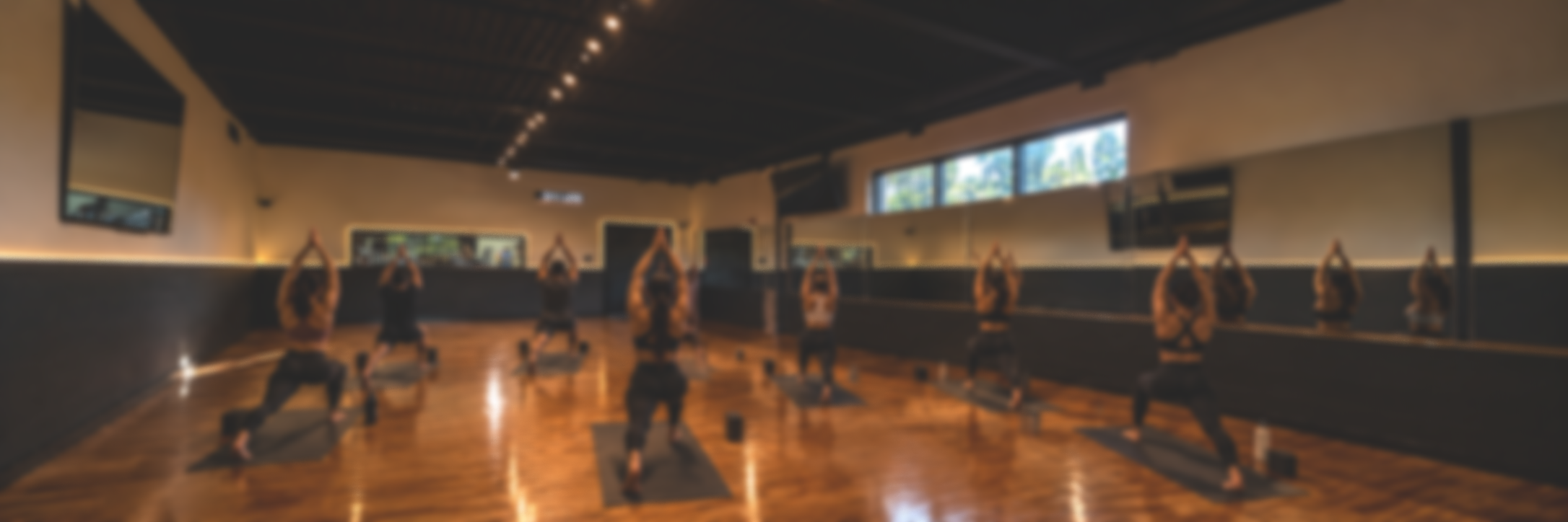 A group of people practicing yoga in a studio, all in the same pose with arms raised overhead, on mats, in a spacious room with wood floors and large windows.