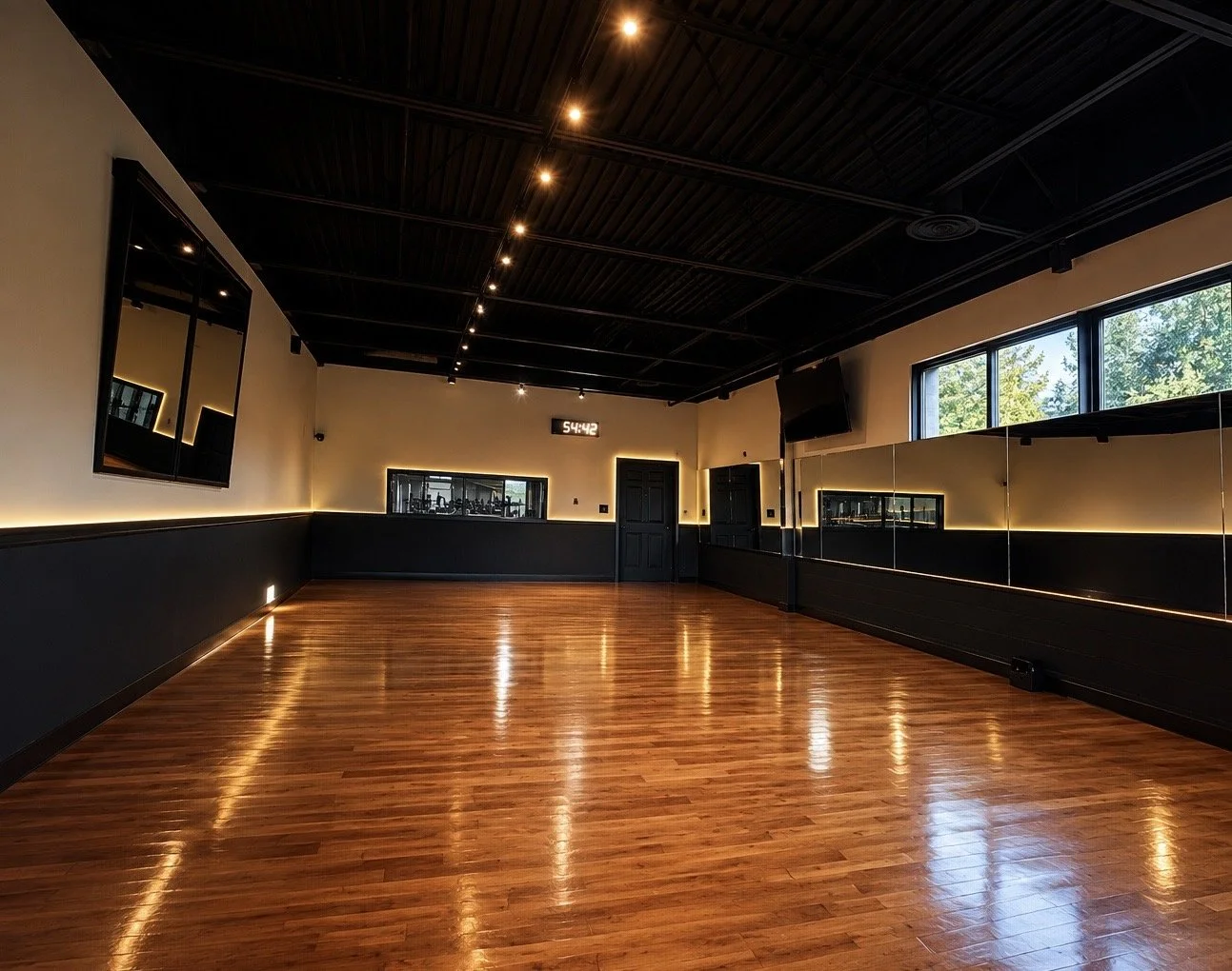 Empty dance studio with hardwood floor, black ceiling, large windows, wall mirrors, and LED lighting.