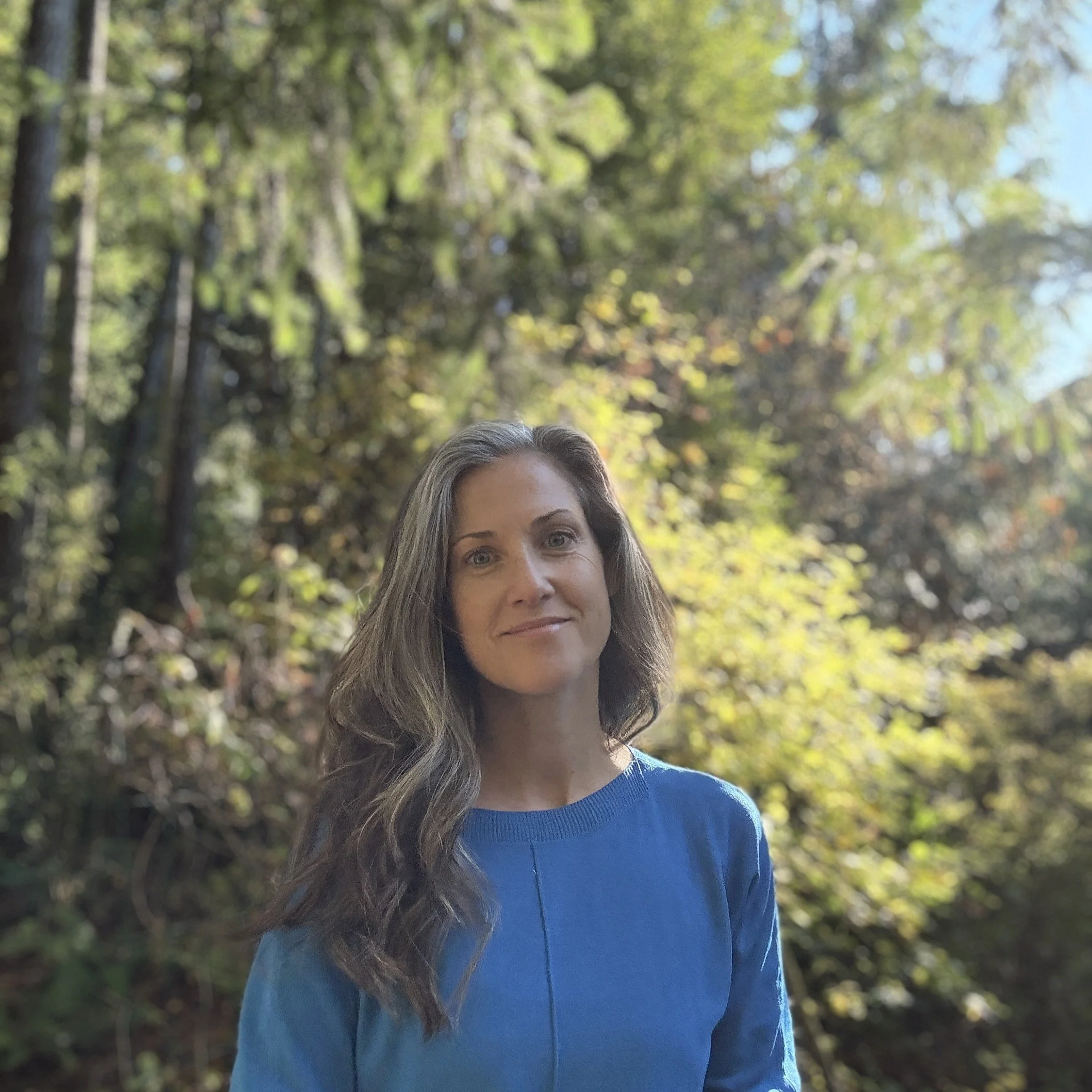 A photo of Ruth, a middle-aged woman with long brown and grey hair, wearing a blue top, outdoors with blurred trees and sunlight in the background.