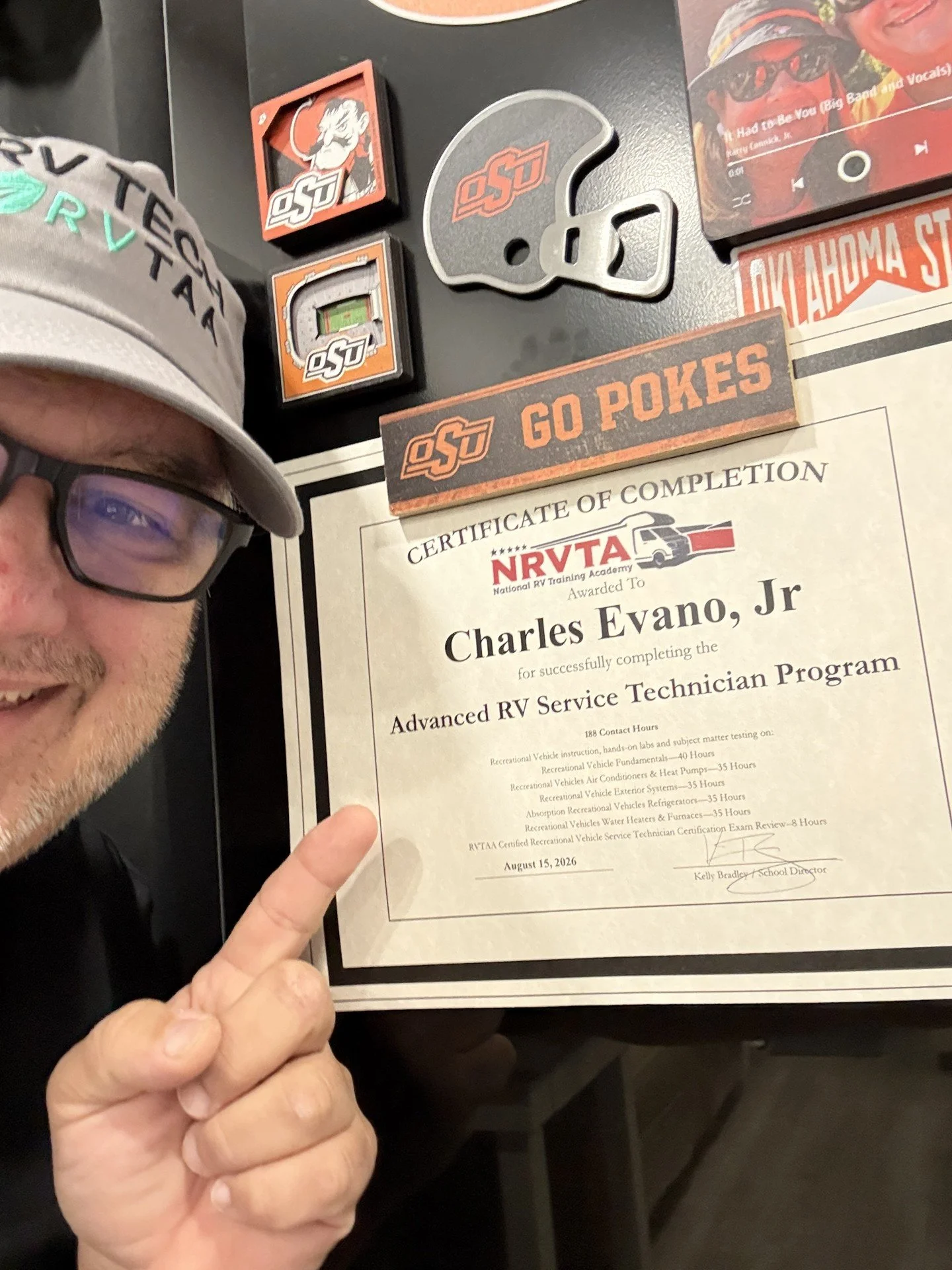 A person in glasses and a cap is holding a certificate of completion from the NRVTA. The certificate is awarded to Charles Evano Jr., for completing the Advanced RV Service Technician Program, dated August 15, 2026. The background includes Oklahoma State University memorabilia and a signed football helmet.