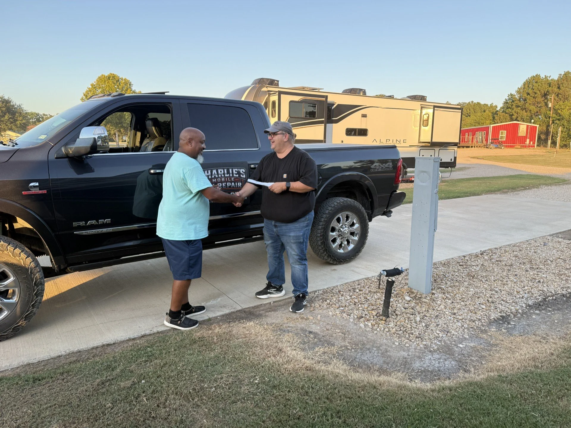 Two men shaking hands outside a black RAM pickup truck with a large RV or camper trailer in the background. One man is bald with a beard, wearing a light blue shirt and dark shorts. The other man is wearing glasses, a gray cap, a black T-shirt, and jeans. The setting appears to be a parking area with a clear sky.