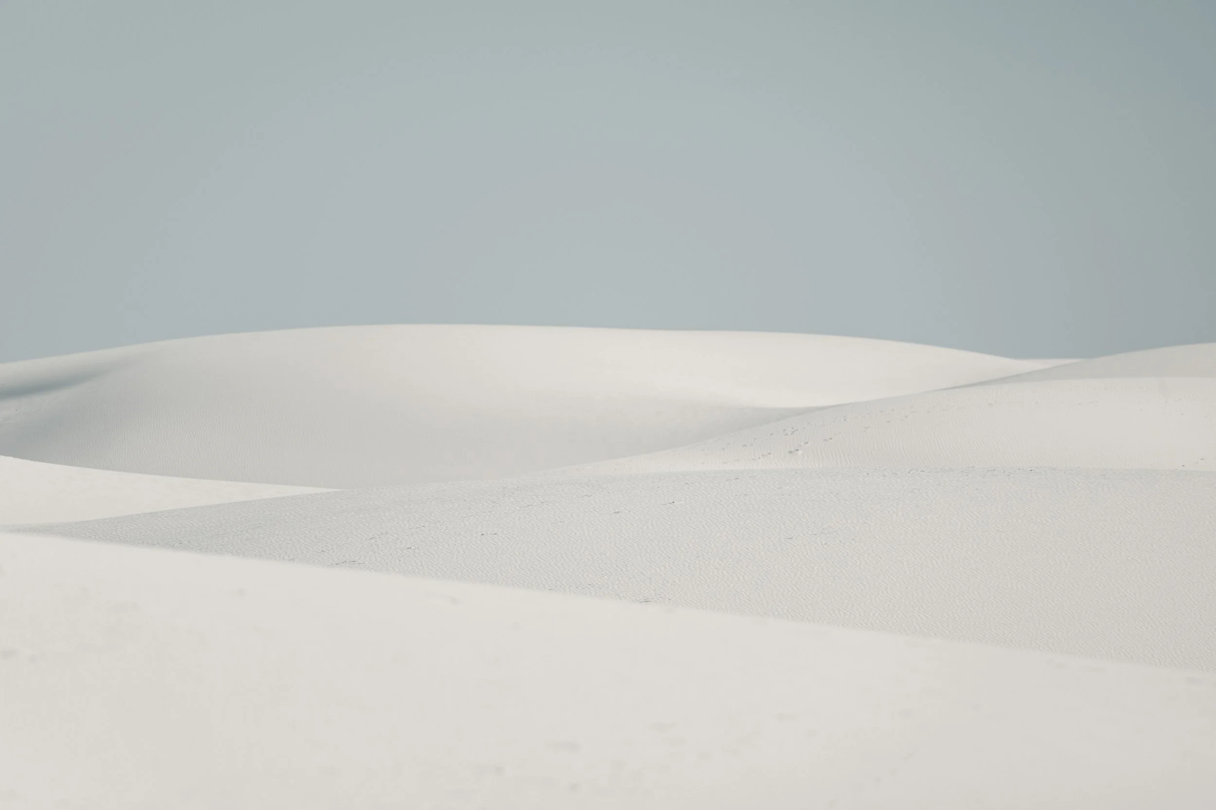 Blank white sandy desert dunes under a pale blue sky with no people or objects visible.