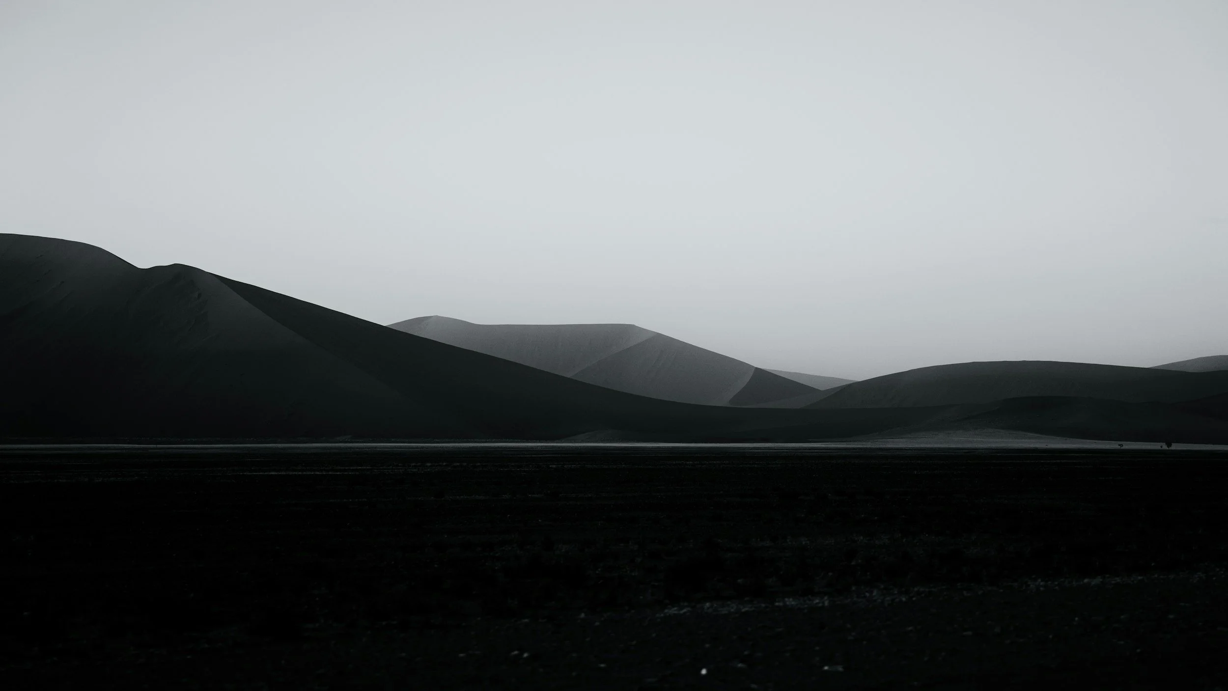 A black and white photograph of mountain ranges with smooth, rolling hills and a flat foreground, under a clear sky.