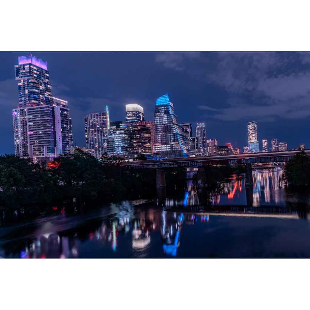 Austin skyline at night mirrored in Lady Bird Lake, with illuminated downtown towers and a railroad bridge