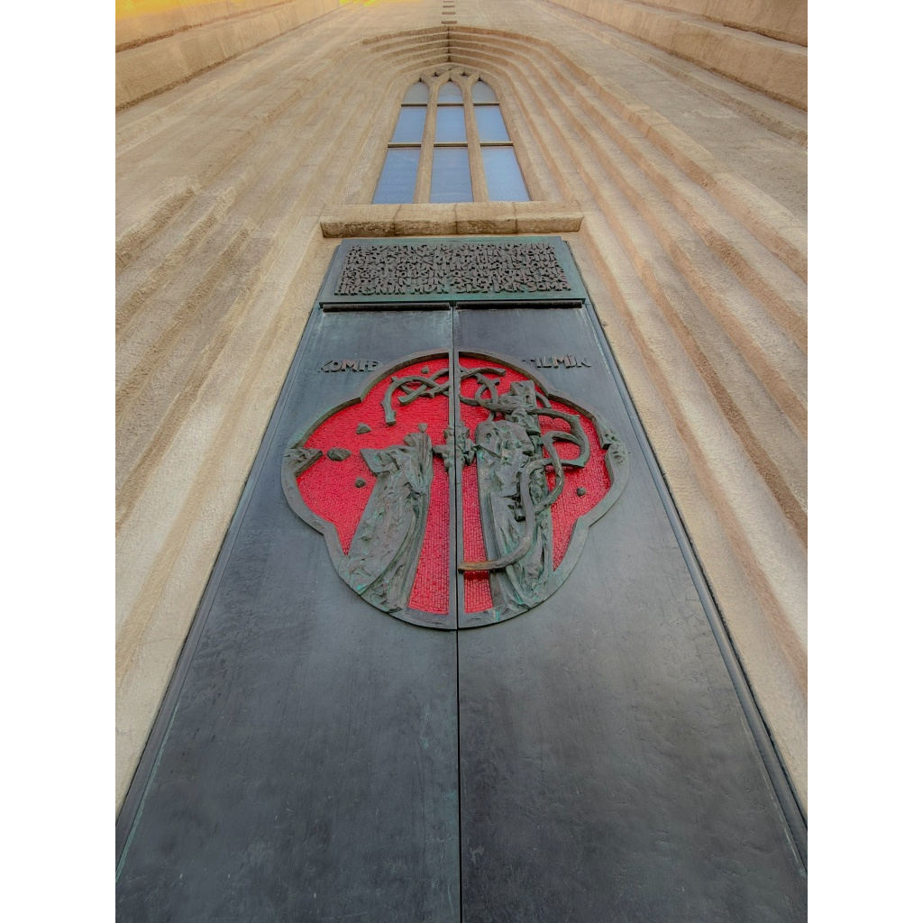 Low-angle view of Hallgrímskirkja’s dark metal doors with red medallion and “KOMIÐ TIL MÍN” lettering