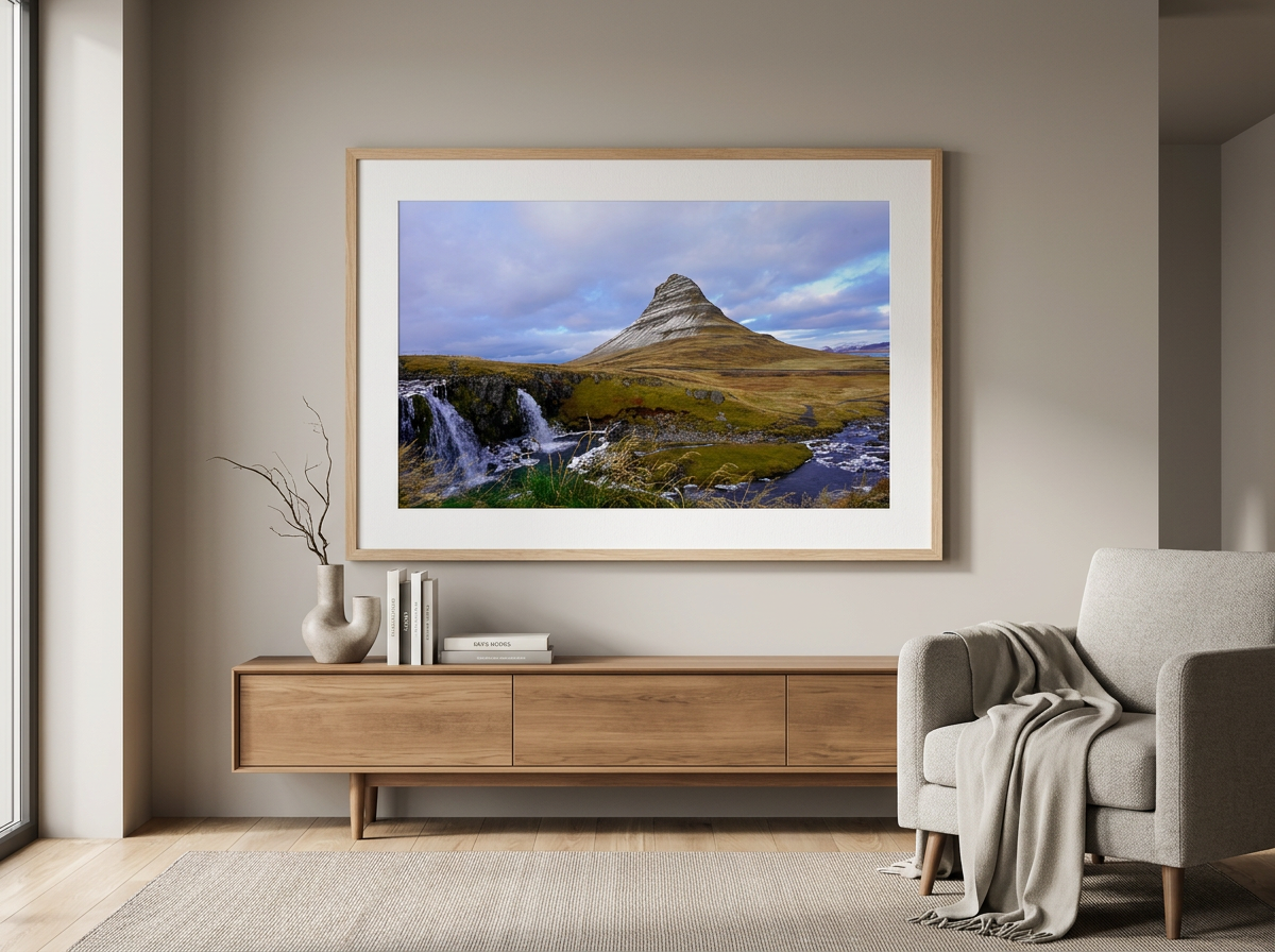 Living room with a large framed landscape photograph of a mountain, waterfall, and river hanging on a beige wall. A wooden sideboard below holds a white vase with twigs and a few books. A gray armchair with a throw blanket is to the right, and a window with sunlight illuminates the room.