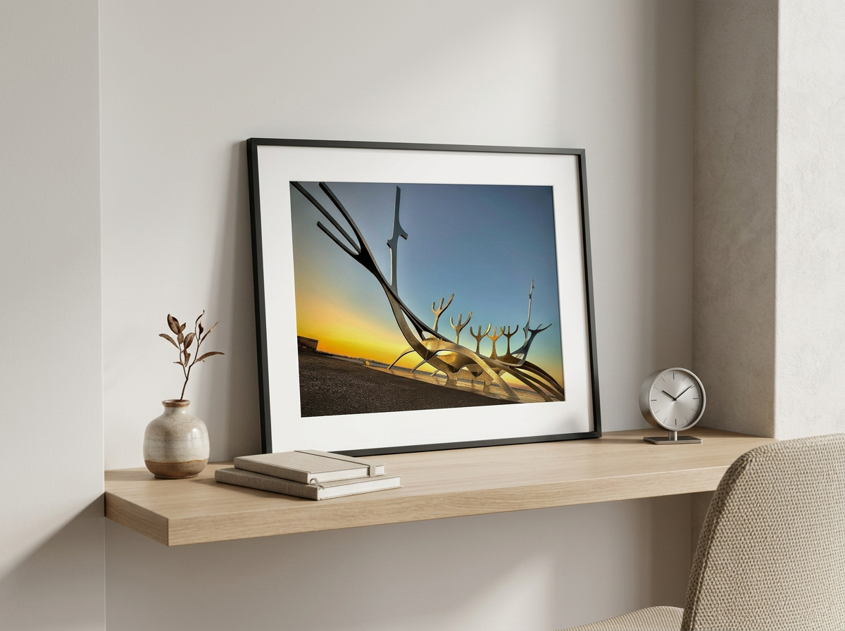 Contemporary art sculpture of a ship's bow with elongated, curved elements, set against a sunset sky. The sculpture leans on a white wall, on a wooden desk with a small vase, books, and a round clock.