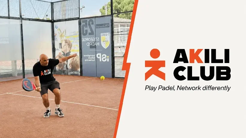 Entrenador o jugador en una cancha de pádel en posición de juego, sosteniendo una raqueta y usando ropa deportiva, con un fondo de paredes de malla y anuncios publicitarios. Parte derecha del imagen con logotipo y texto de AKILI CLUB con lema 'Juega pádel, conecta diferente'.