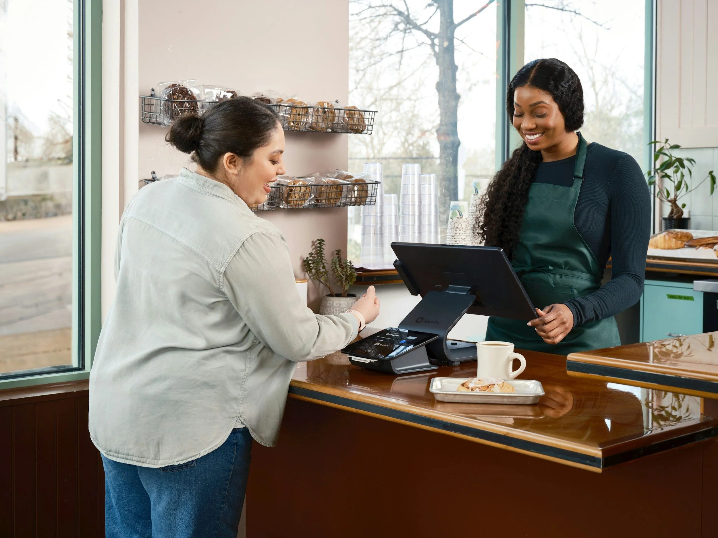 A woman in a white shirt and jeans is paying at a cafe counter using a credit card. Another woman wearing a green apron and dark shirt is smiling at her and working on a point-of-sale terminal. There are cookies in plastic bags on wire shelves on the wall, a window with trees outside, and a tray with pastries and a coffee mug on the counter.