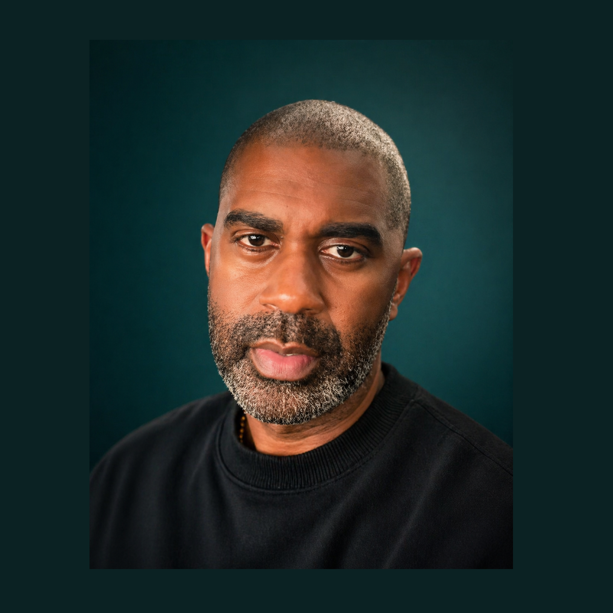 Close-up of a man's face with a well-groomed beard and short hair, wearing a black shirt, against a dark teal background.