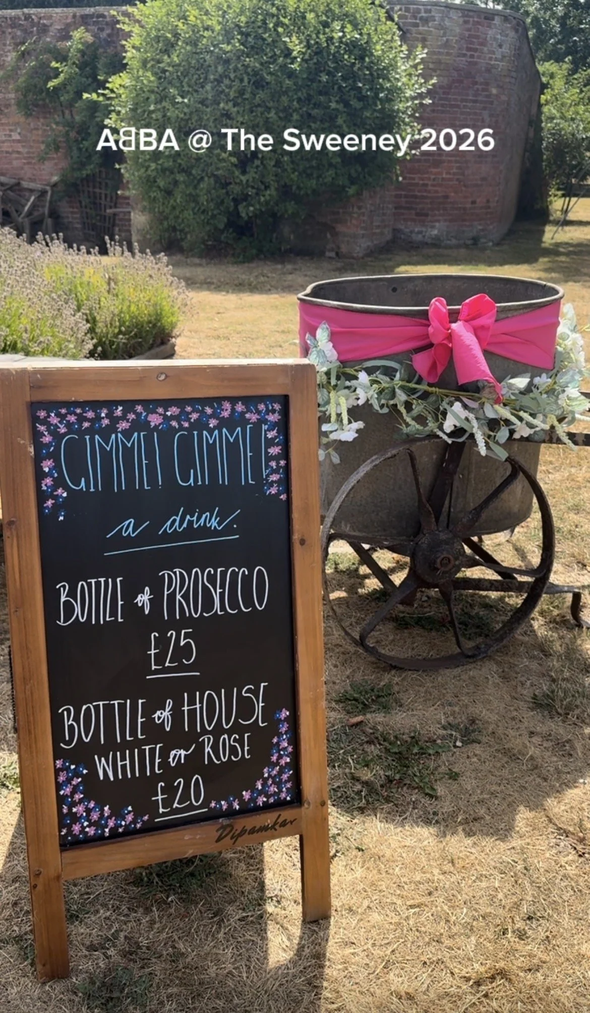 A wooden table with a chalkboard sign advertising drinks, including a bottle of prosecco for £25 and a bottle of house white or rosé for £20. Behind the sign is an old wheelbarrow decorated with pink satin fabric, flowers, and a large pink bow. There are bushes and brick structures in the background, indicating an outdoor setting during the day.