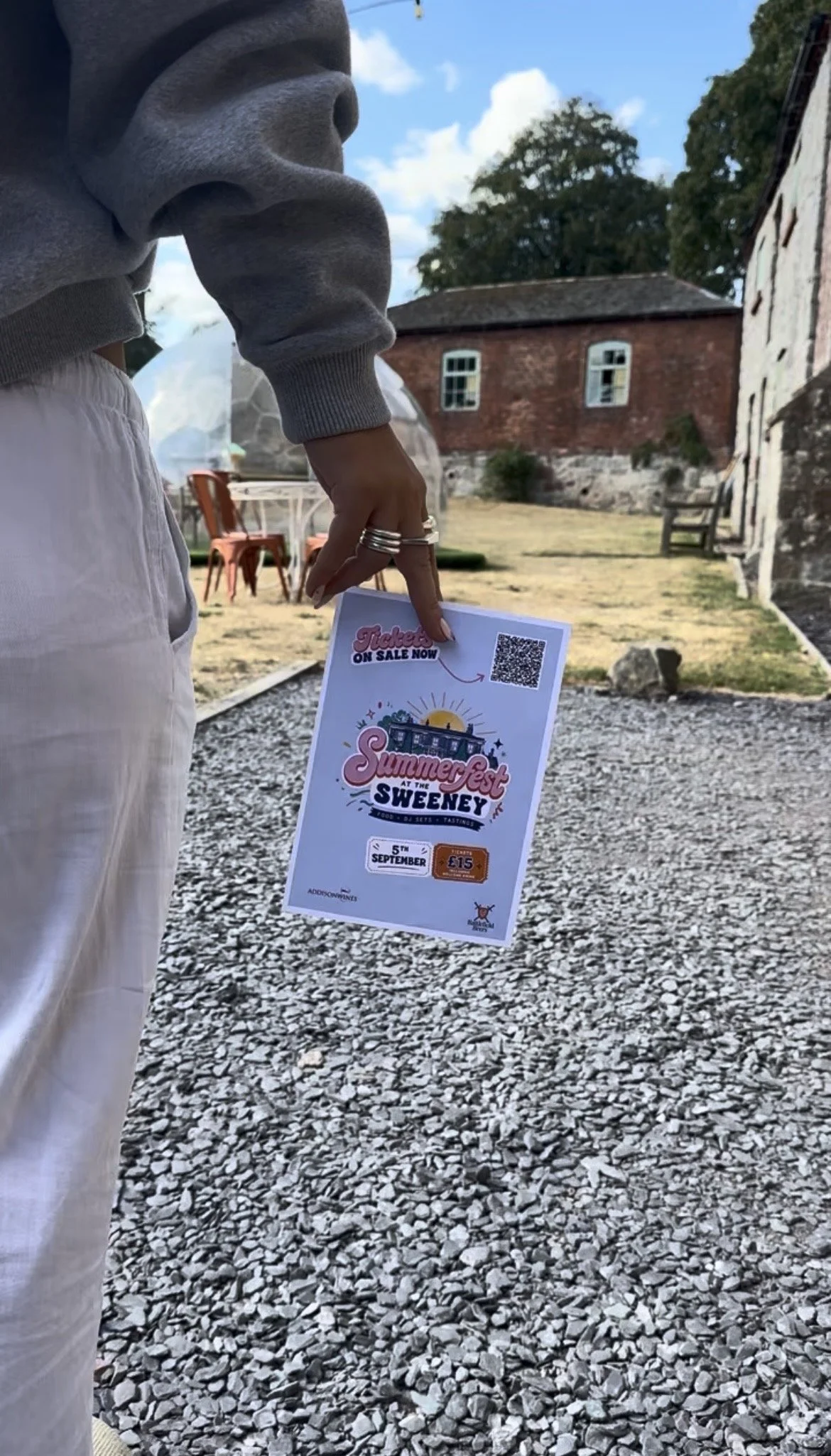 Person holding a flyer for Summer Fest at the Sweeney, scheduled for September 5th, with a QR code, ticket price, and promotional graphics, outdoors with a background of old buildings and trees.