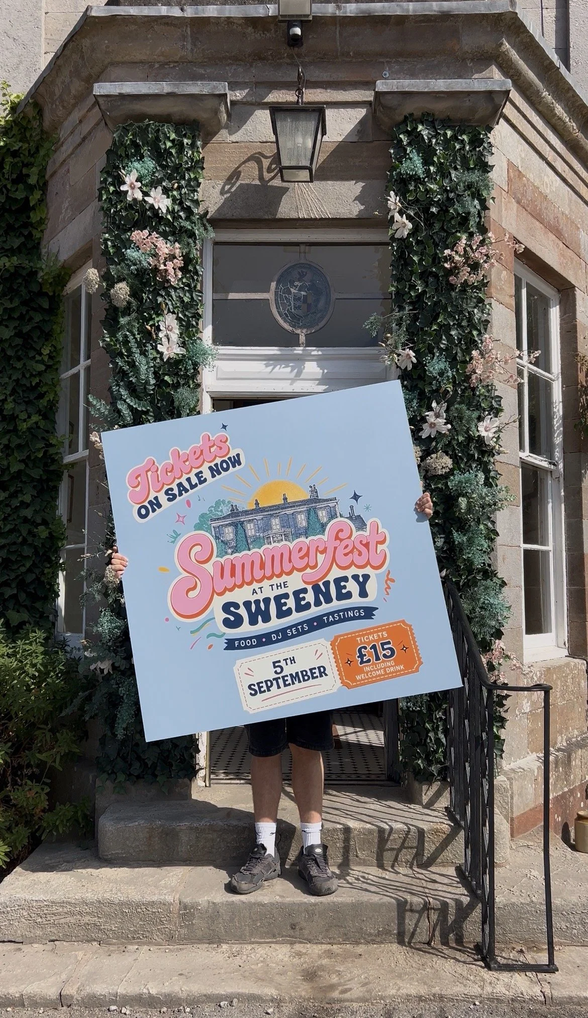 Person holding a large colorful event poster in front of a stone building decorated with flowers and greenery. The poster advertises summer event details including date, ticket price, and activities.