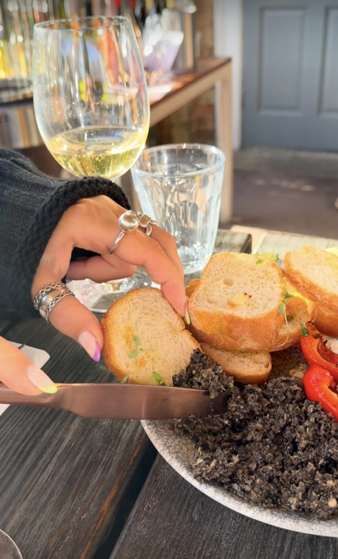 A person's hand with rings cutting bread on a plate of food. The plate also contains black rice, cherry tomatoes, and garnishes. There are glasses of white wine and water on the table, with a blurred background of a rustic setting.