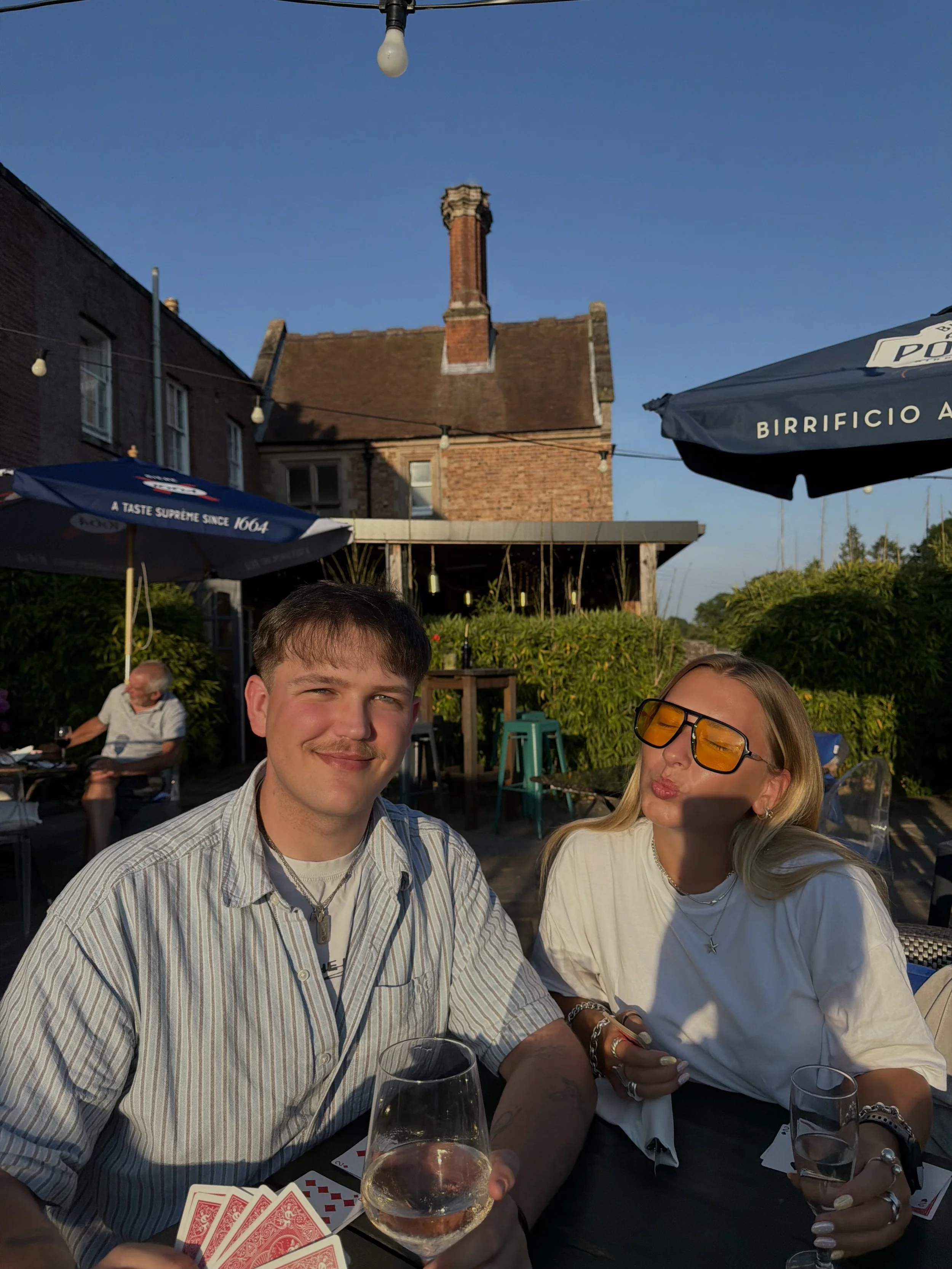 Two people sitting at an outdoor table during sunset, with a man holding a glass of white wine and a woman wearing yellow-tinted sunglasses. There are playing cards on the table, and a background of brick buildings with a clear blue sky and string lights.
